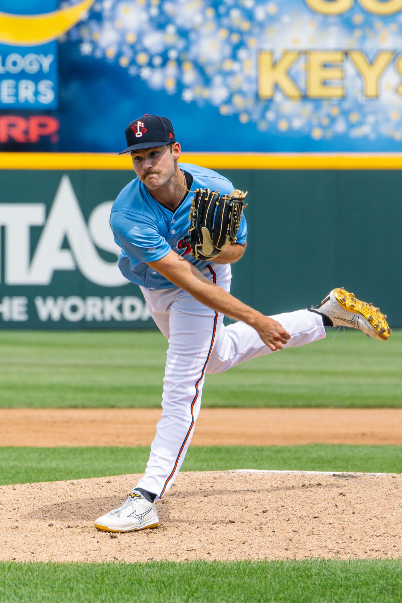 A baseball pitcher in a light blue uniform mid-windup on the mound during a sunny game.