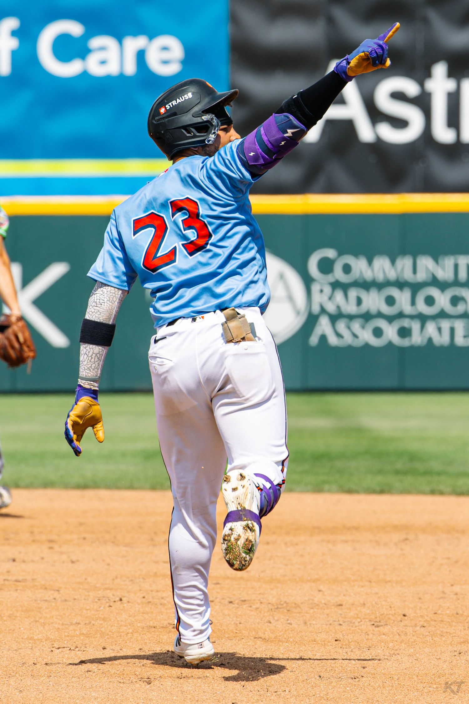 Baseball player #23 in blue uniform celebrates rounding bases with finger pointed skyward on sunny game day.
