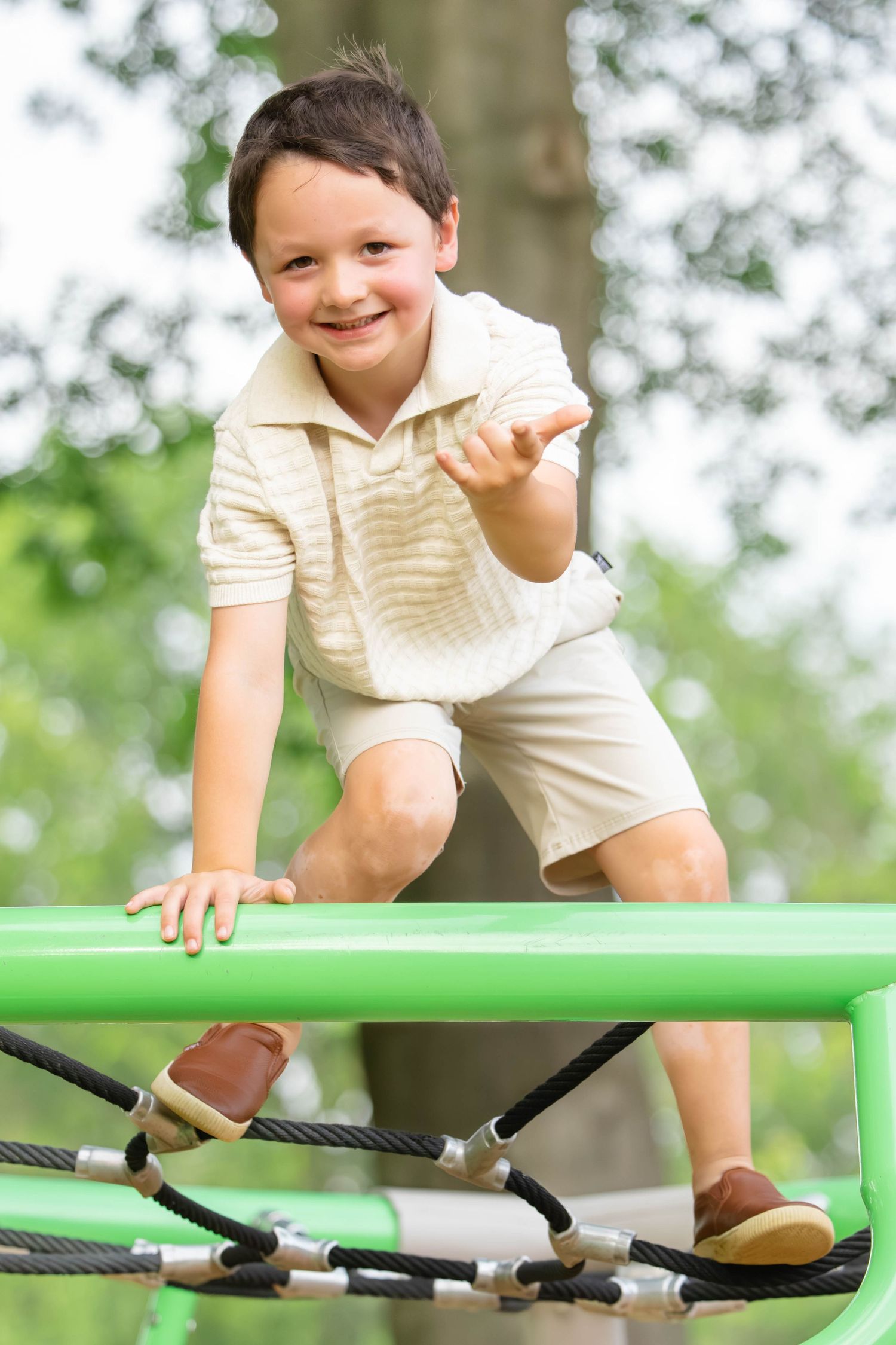 Happy boy climbing on green playground equipment outdoors, smiling and gesturing playfully in a park setting.