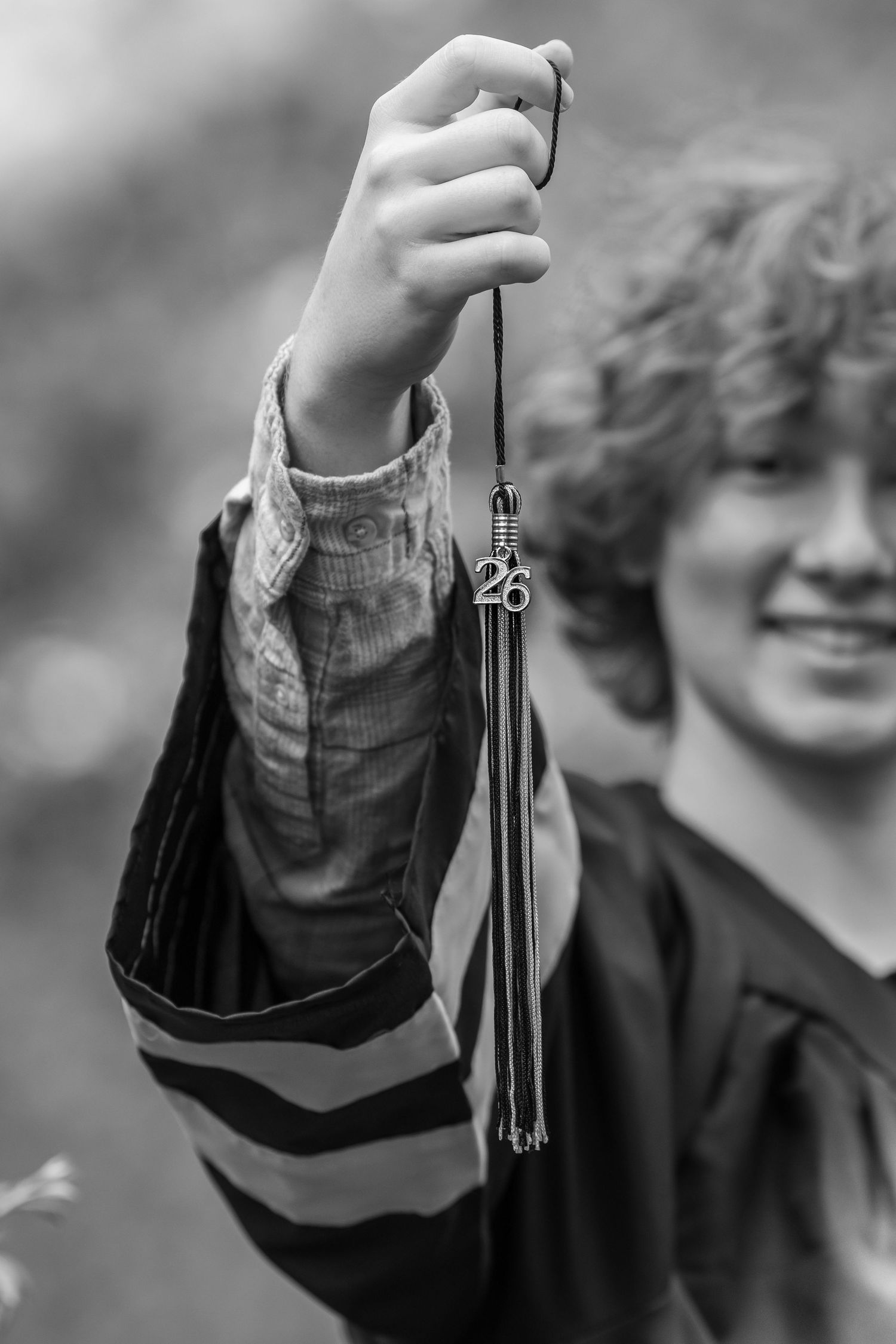 Black and white photo of smiling graduate holding up mortarboard tassel with class of 26 charm.