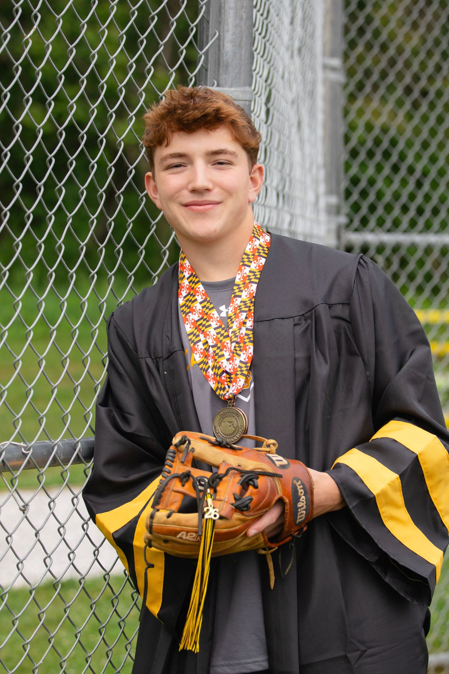 Teen boy in graduation gown holding baseball glove, wearing candy lei and medal, smiling by chain-link fence.
