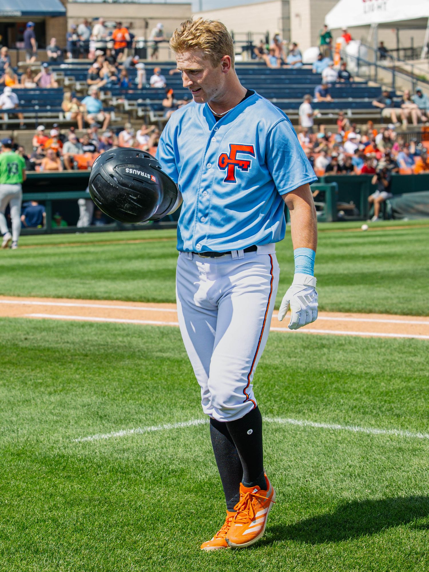 Baseball player in light blue CF uniform walks on field holding helmet during a sunny minor league game.