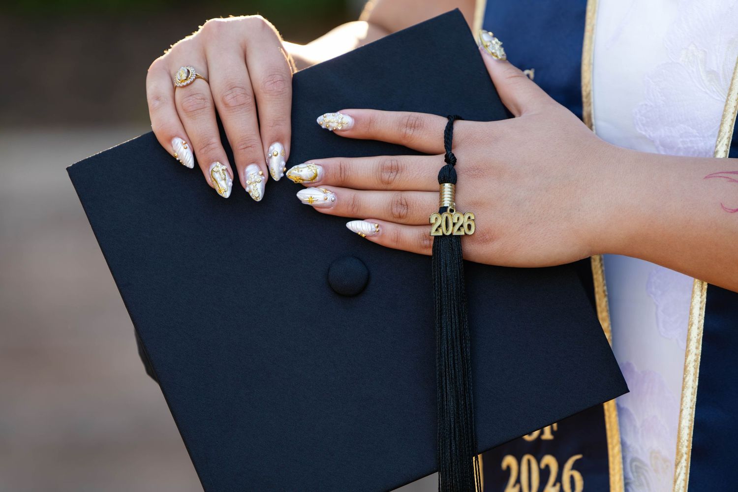 Close-up of decorated graduation cap held by hands with ornate nail art and a 2026 tassel charm.