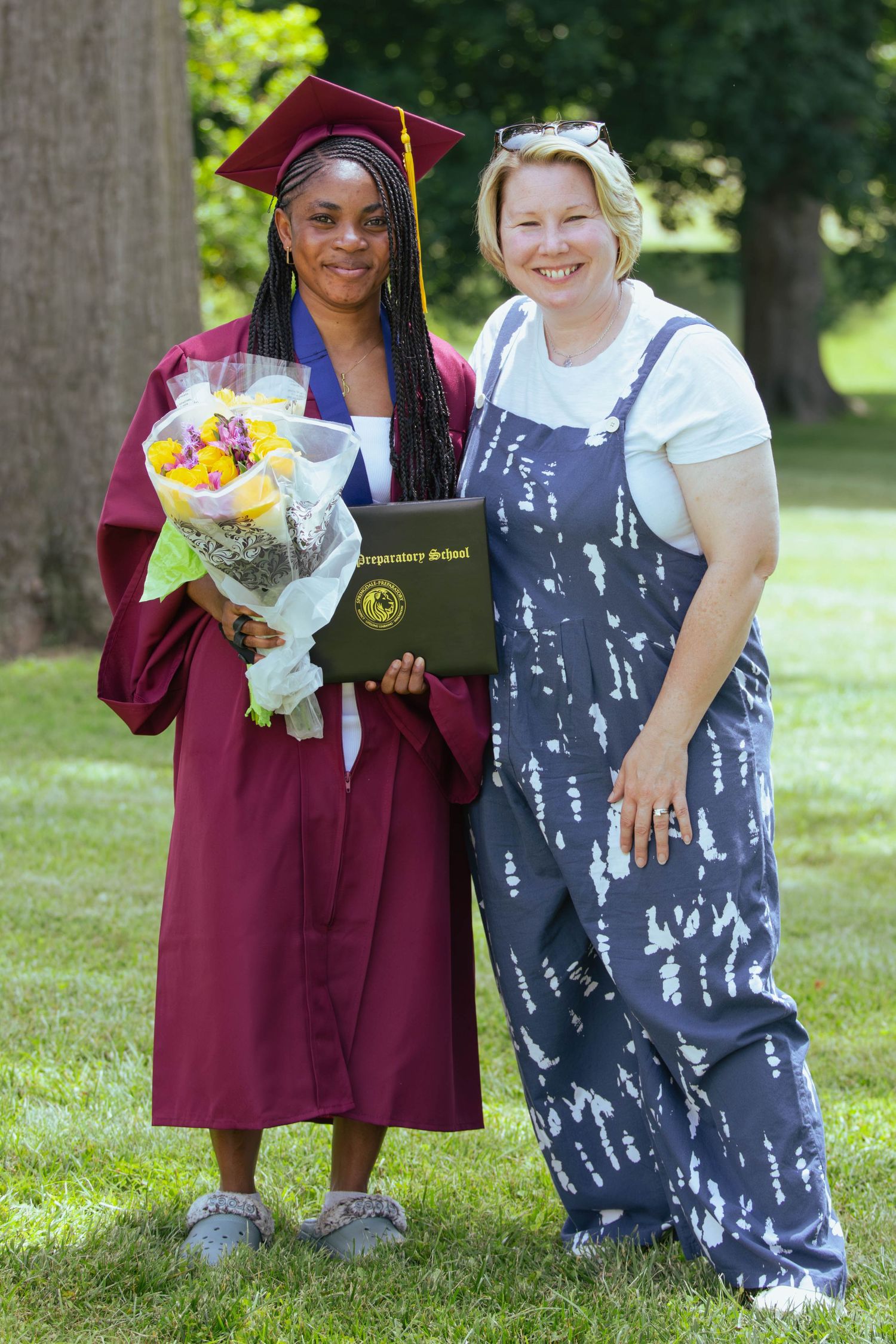 Graduate in maroon cap and gown holds flowers and diploma beside smiling woman in patterned overalls outdoors.