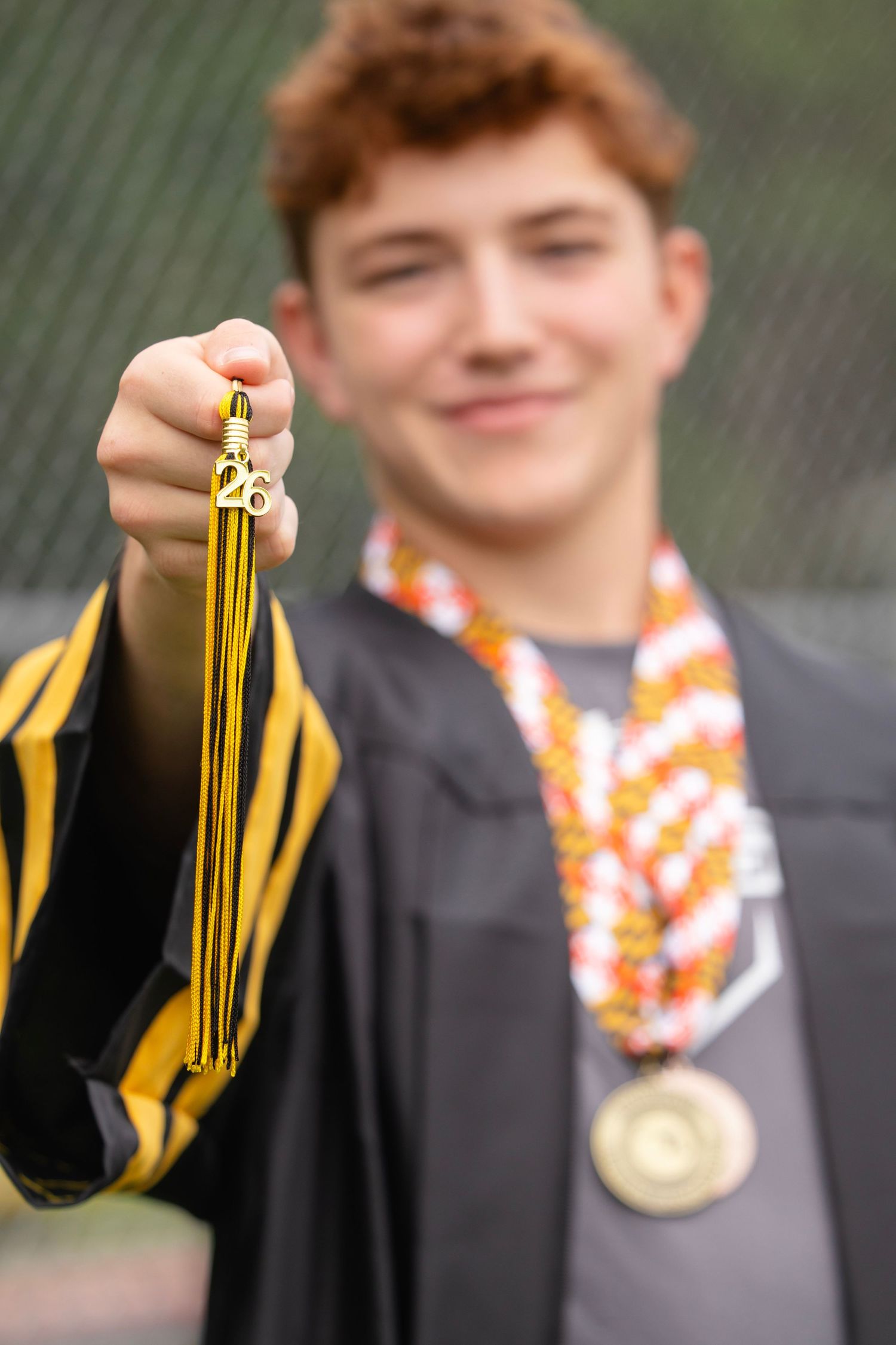 A smiling graduate in black and gold gown holds up a gold tassel while wearing colorful leis and a medal.