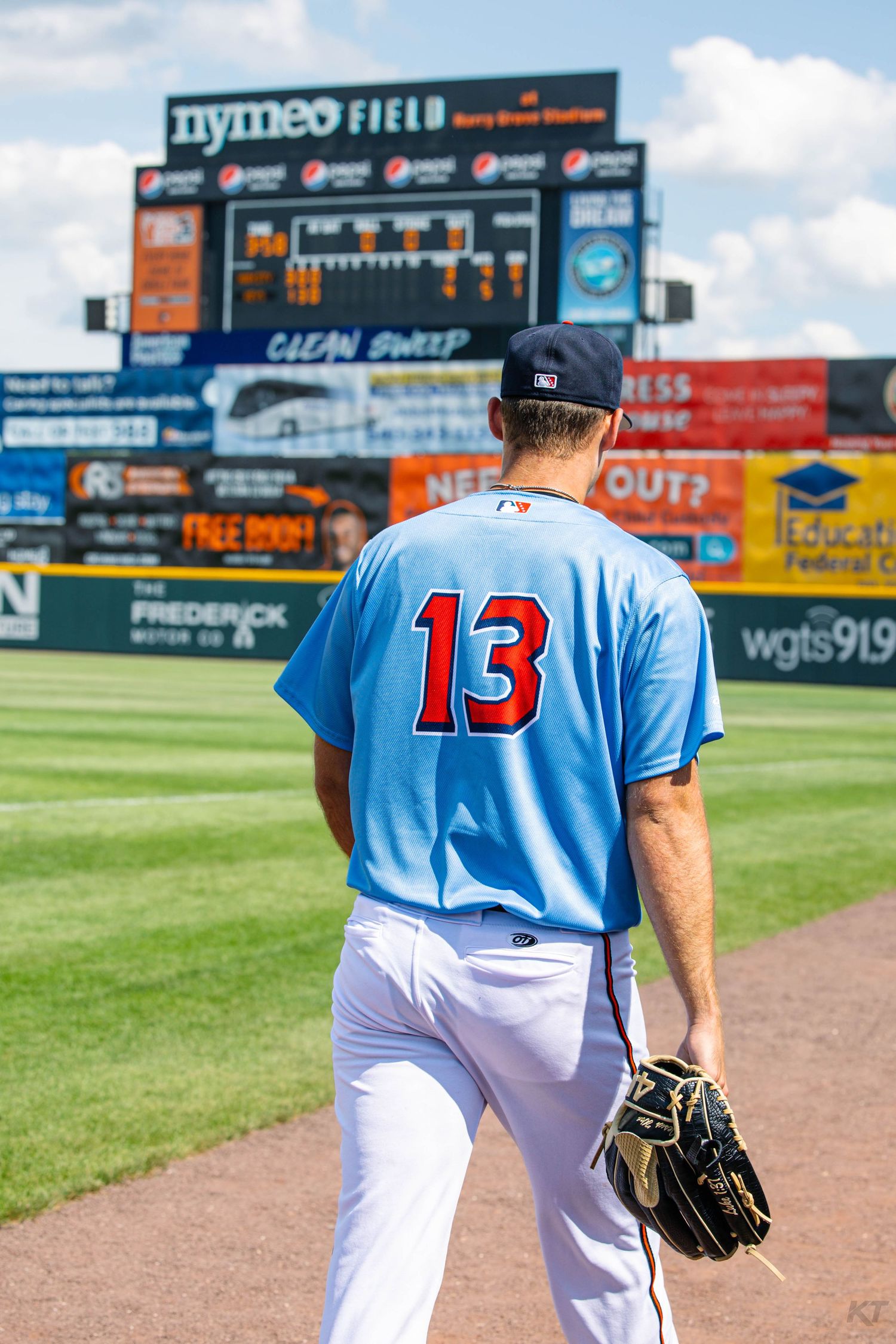 Minor league baseball player wearing number 13 light blue jersey walks toward the scoreboard at Nymeo Field.