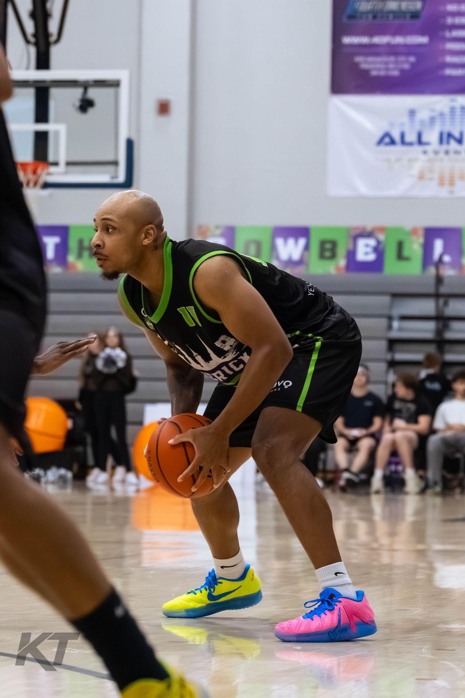 Bald basketball player in black and green jersey dribbles ball on indoor court wearing colorful Nike sneakers.