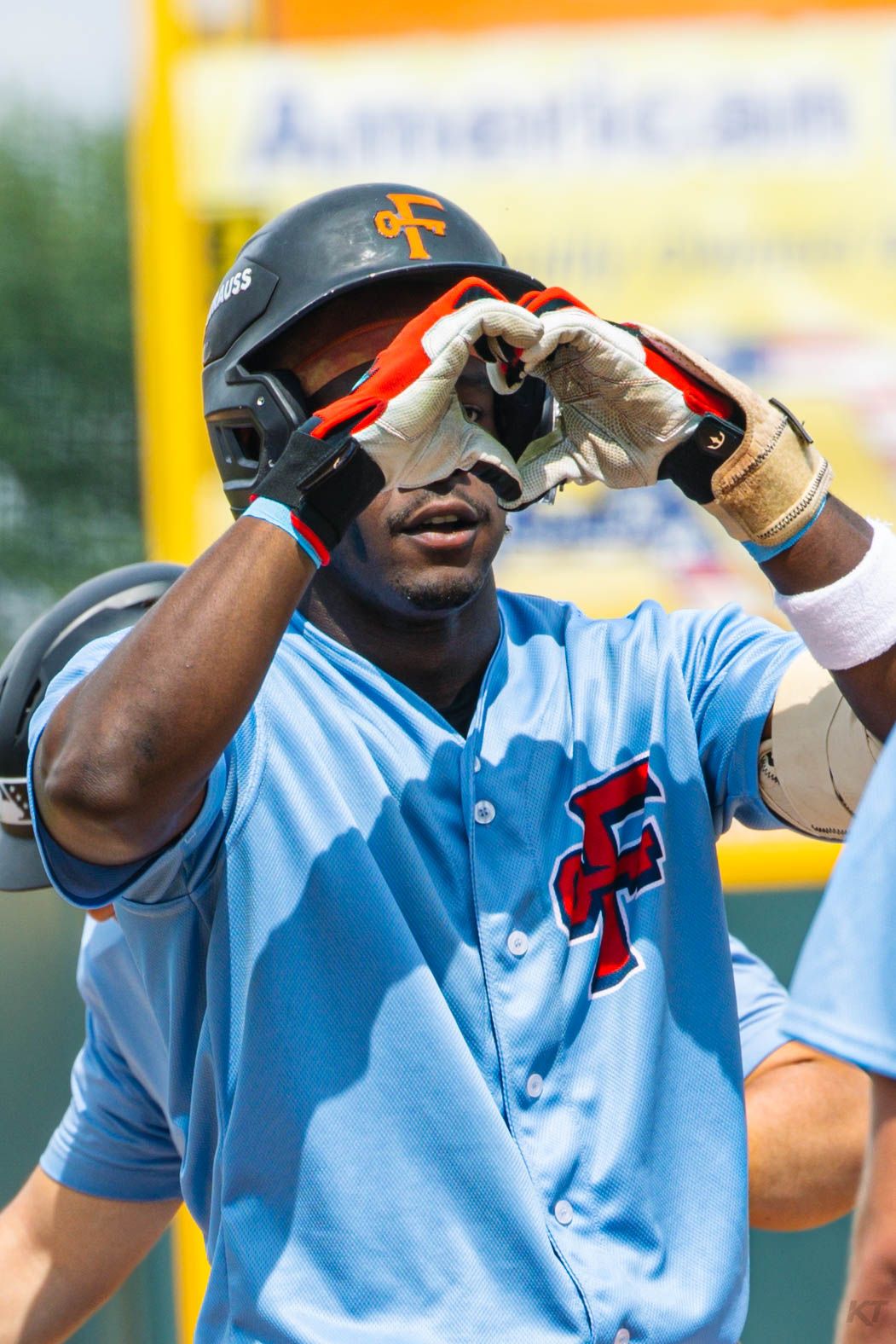 Baseball player in light blue CF jersey and helmet adjusts his batting gloves during a sunny game.
