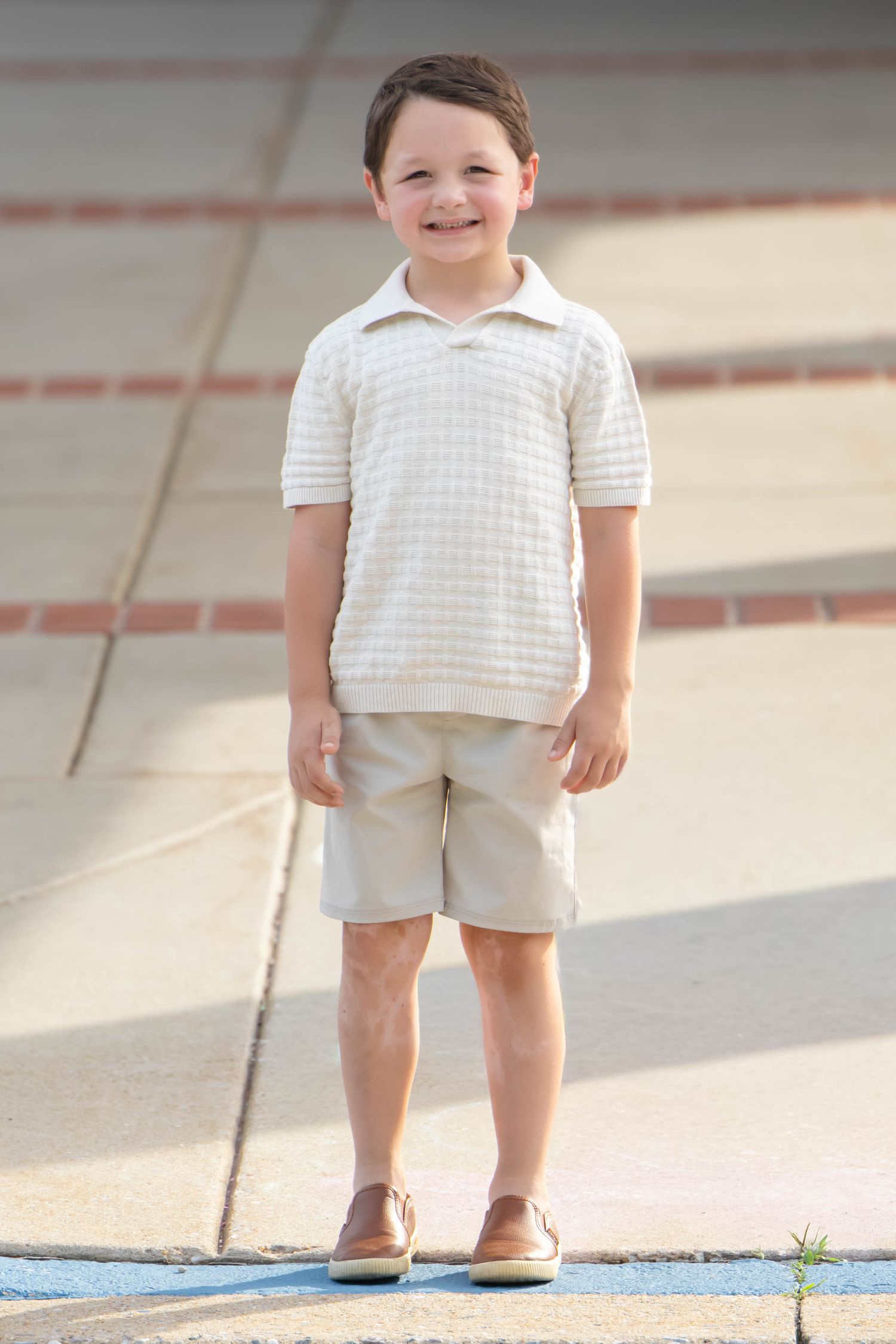 Smiling young boy wearing a white checkered polo shirt and khaki shorts standing outdoors on a sunny day.