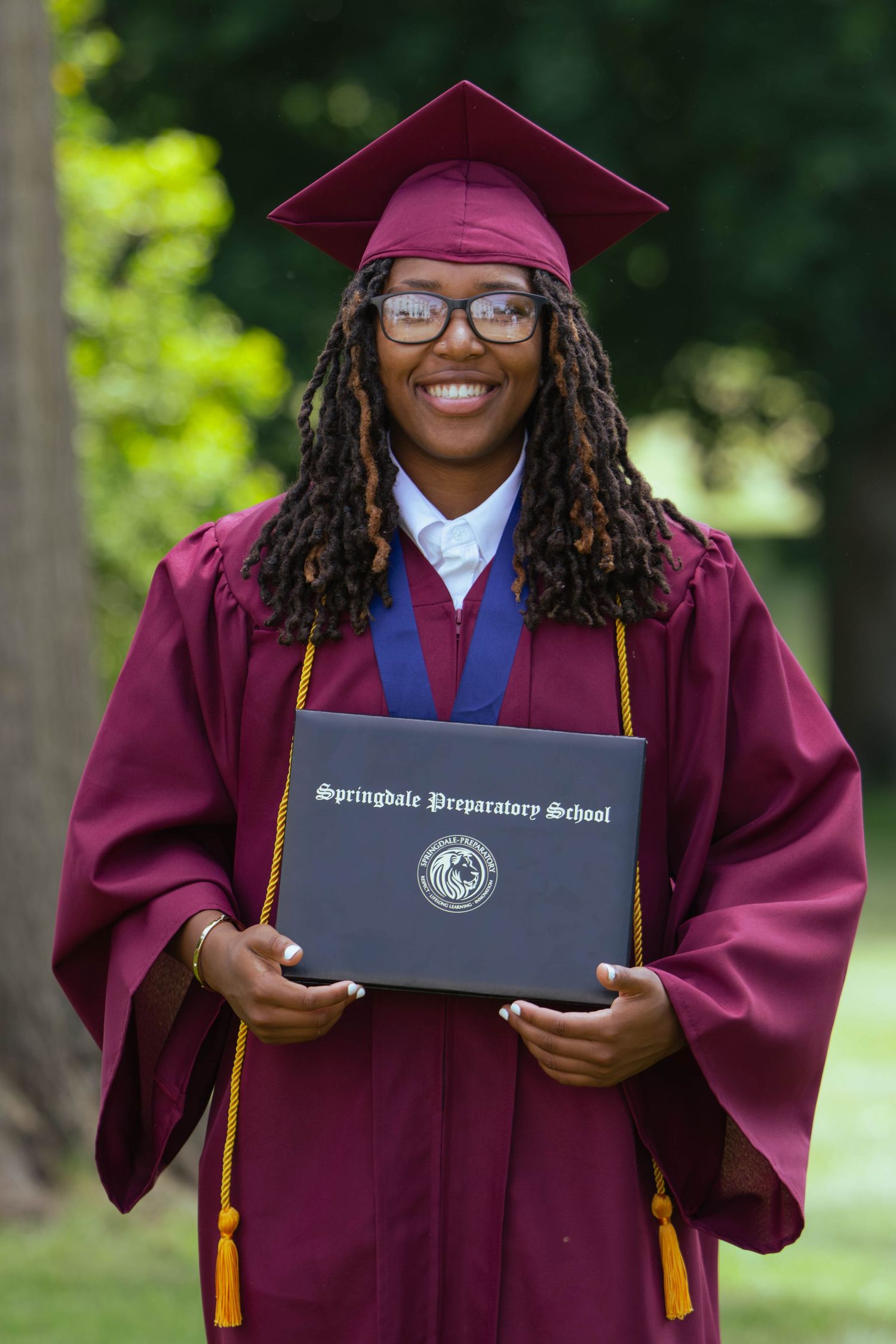 Smiling graduate in maroon cap and gown holds Springdale Preparatory School diploma outdoors.
