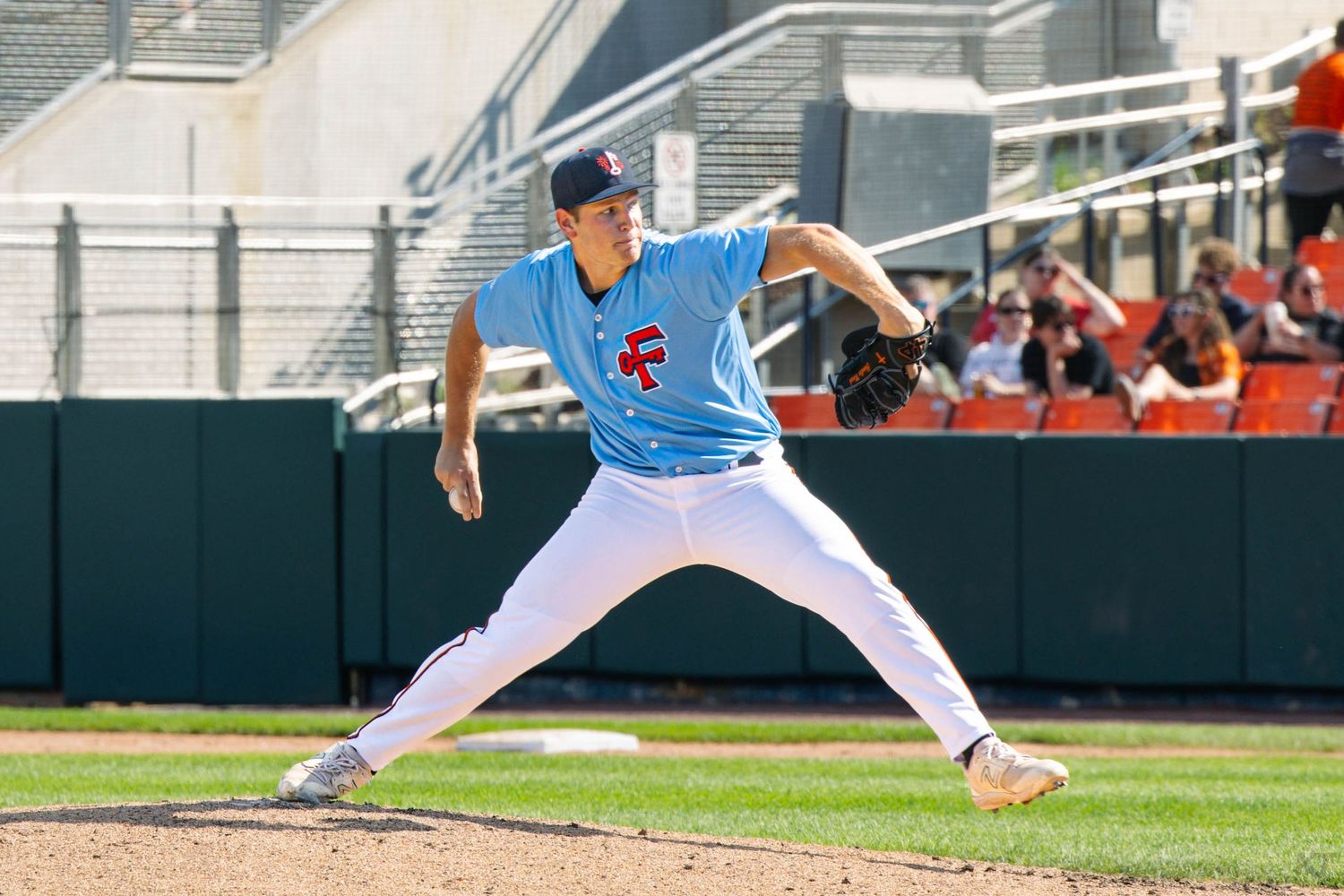 Baseball pitcher in blue uniform mid-wind-up on sunny day at stadium mound.