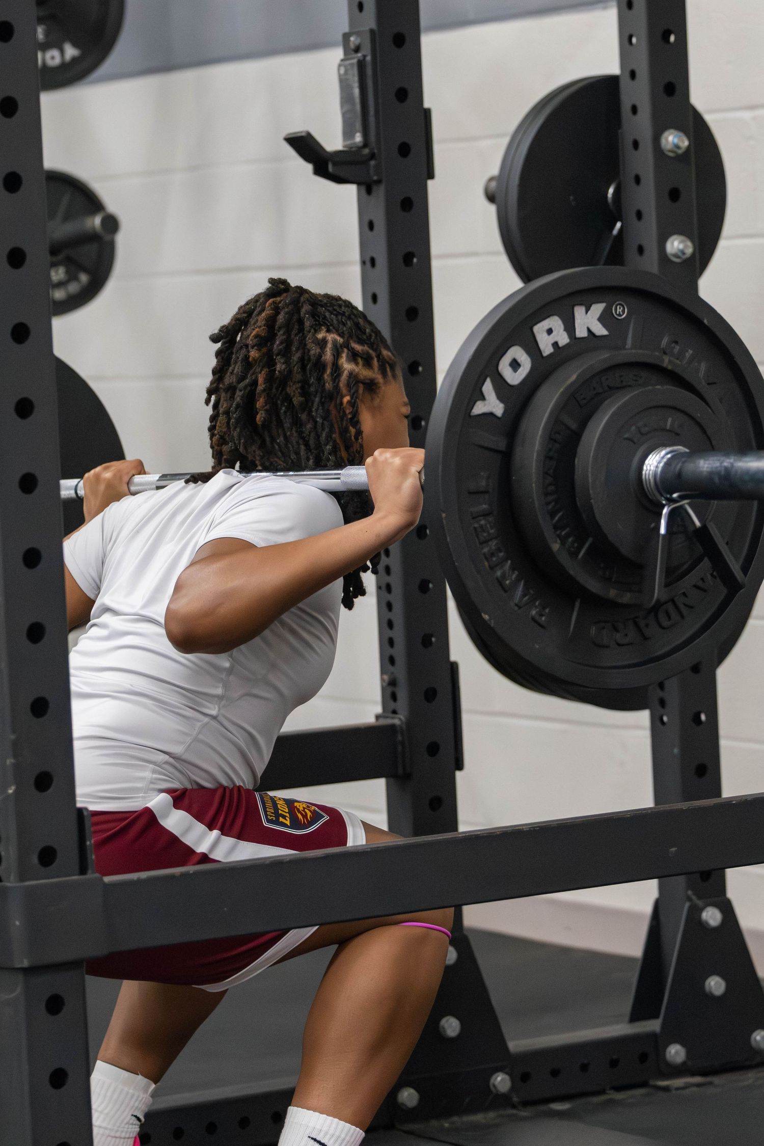 Athlete performing barbell squats on a York weight rack in a gym, wearing white shirt and maroon shorts.