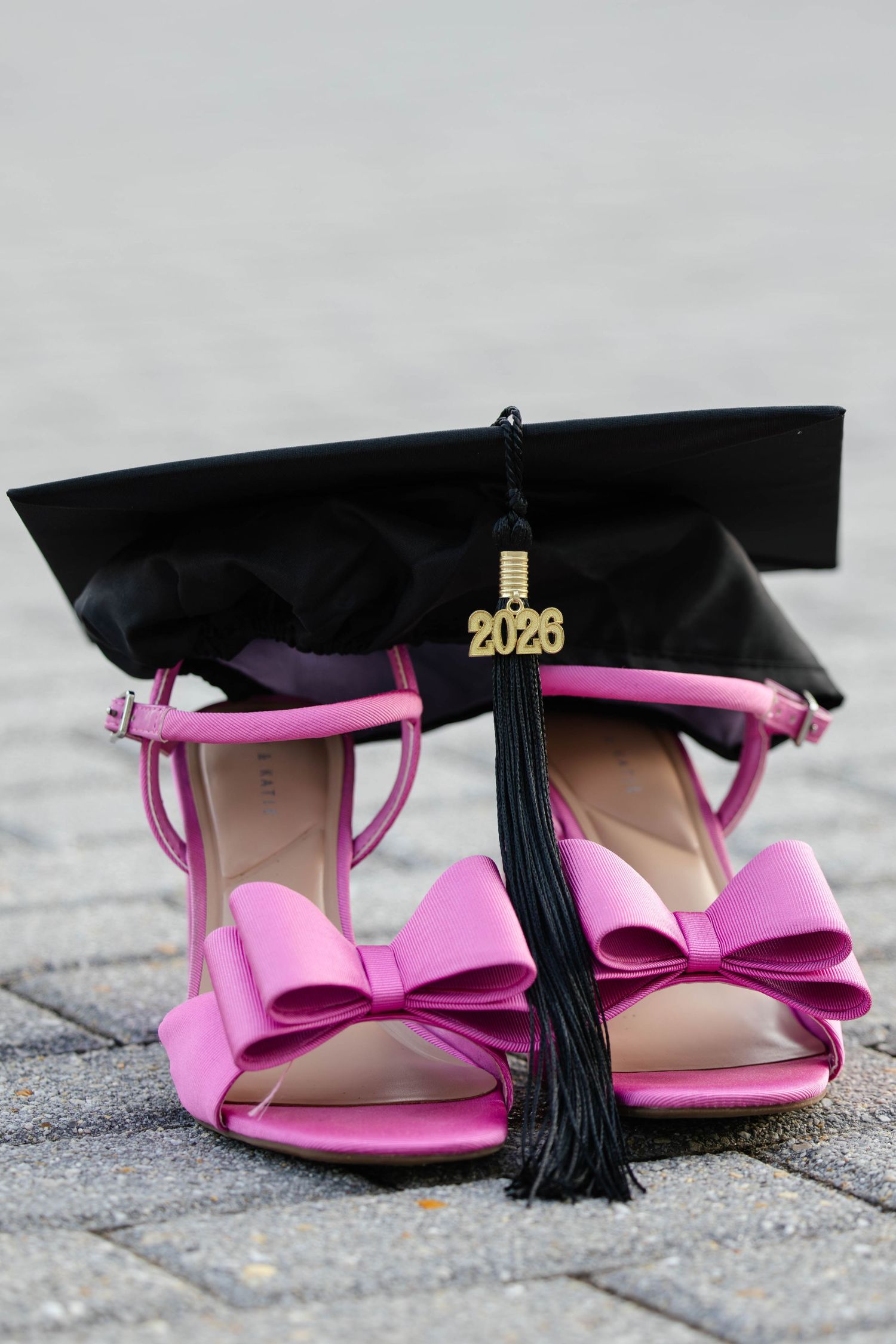 Pink bow heeled sandals with black 2026 graduation cap and tassel on stone pavement.