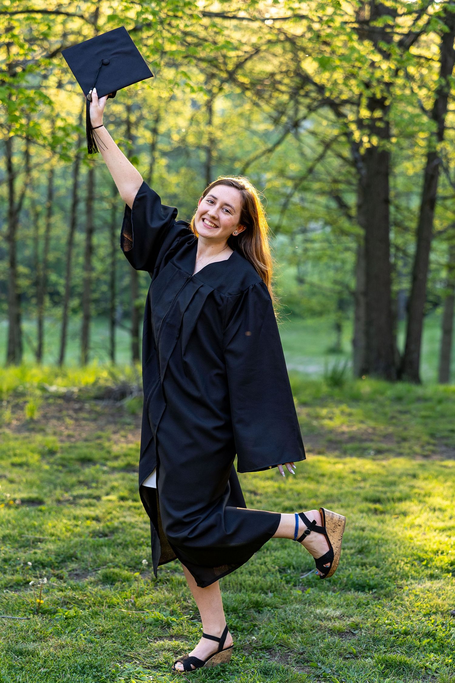 College graduate celebrates outdoors, holding her cap high while wearing a graduation gown in a sunlit park.