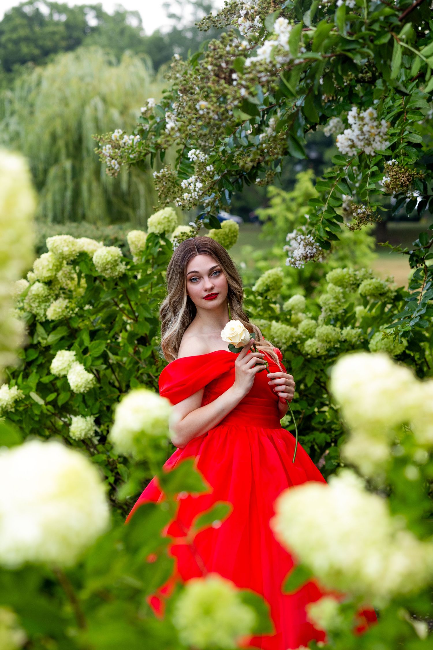 College graduate poses outdoors, holding a white rose while wearing a red ball gown.