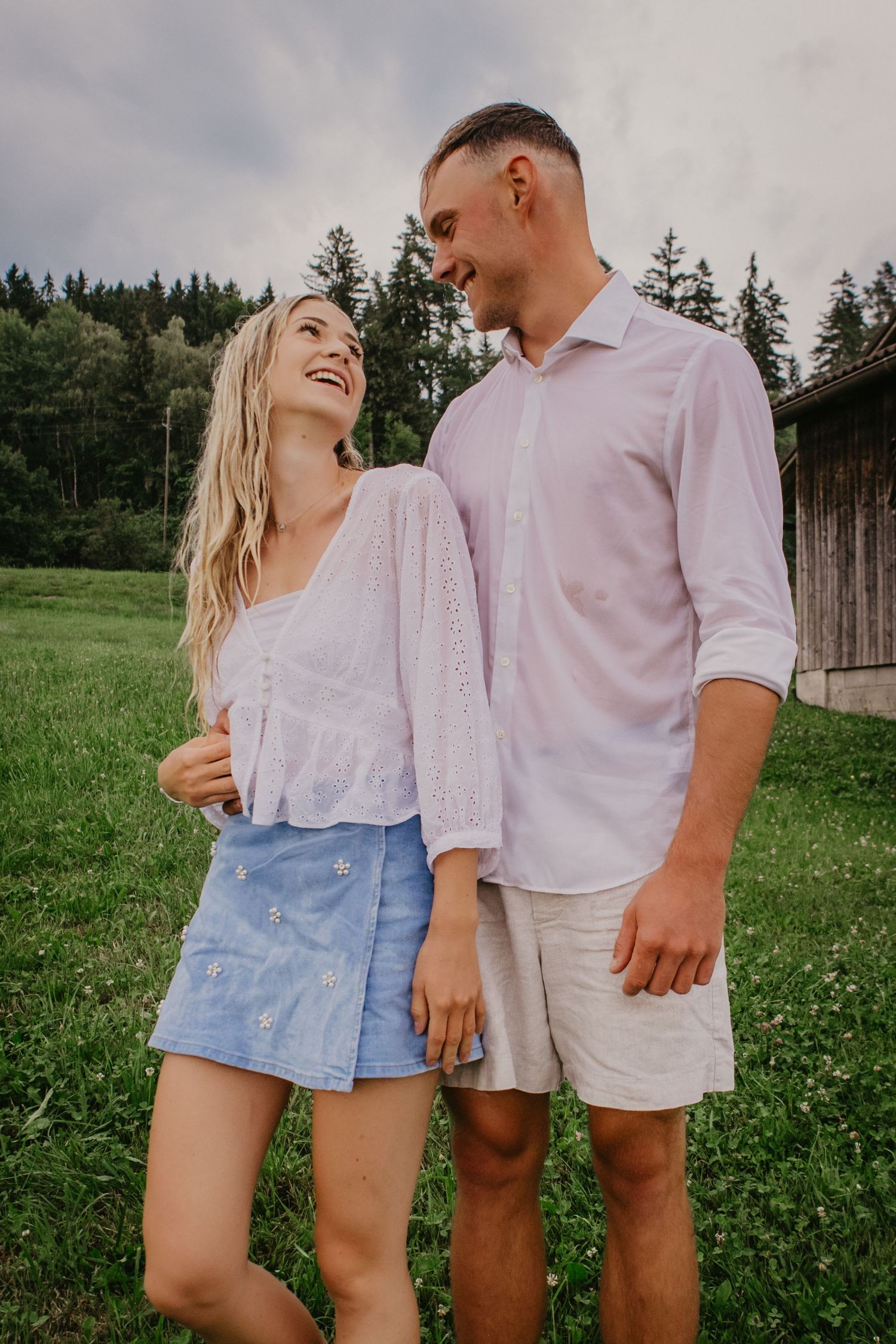Happy couple laughing together outdoors on green grass with forest background, woman in white top and blue skirt.