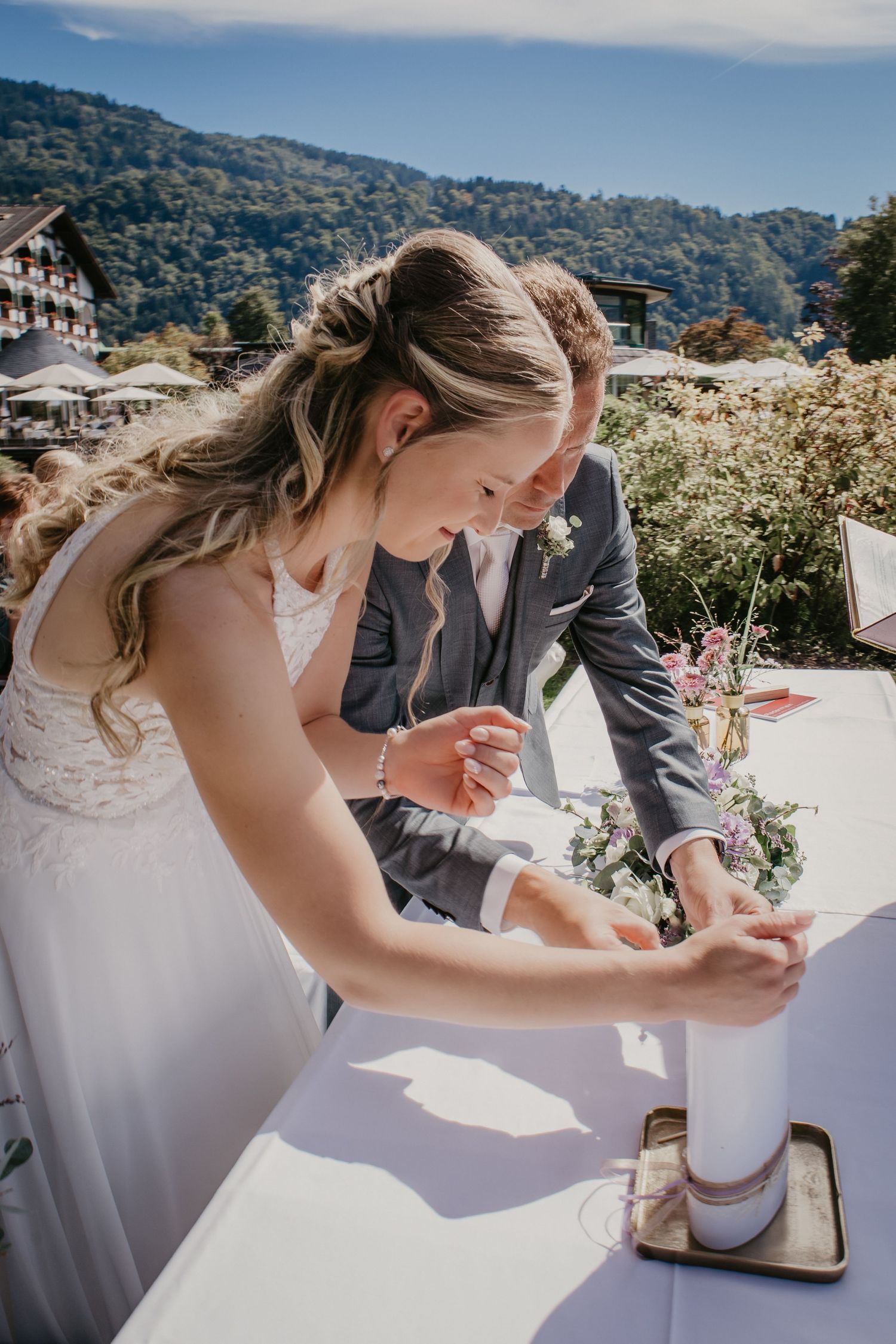 A bride in white lace dress signs wedding documents at an outdoor ceremony.