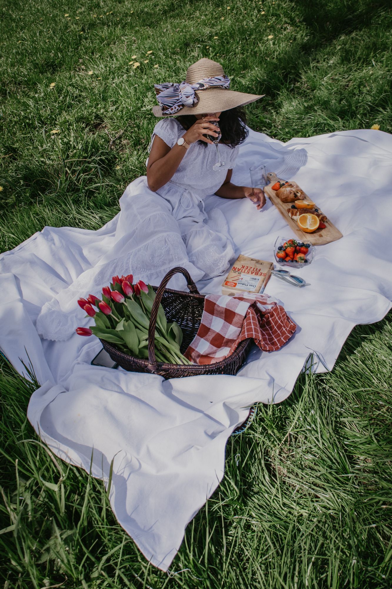 White picnic blanket with food spread and flowers laid out on green grass.