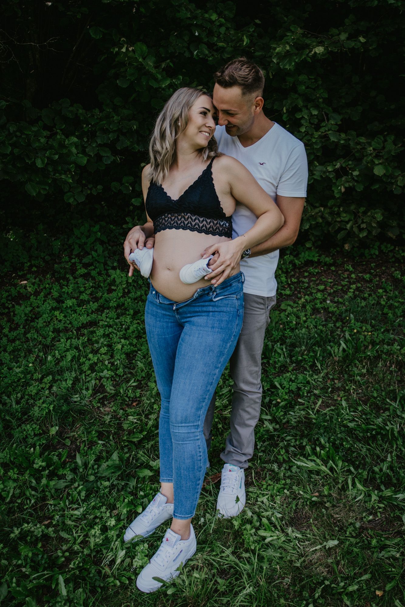 A couple embraces while standing in front of green foliage.