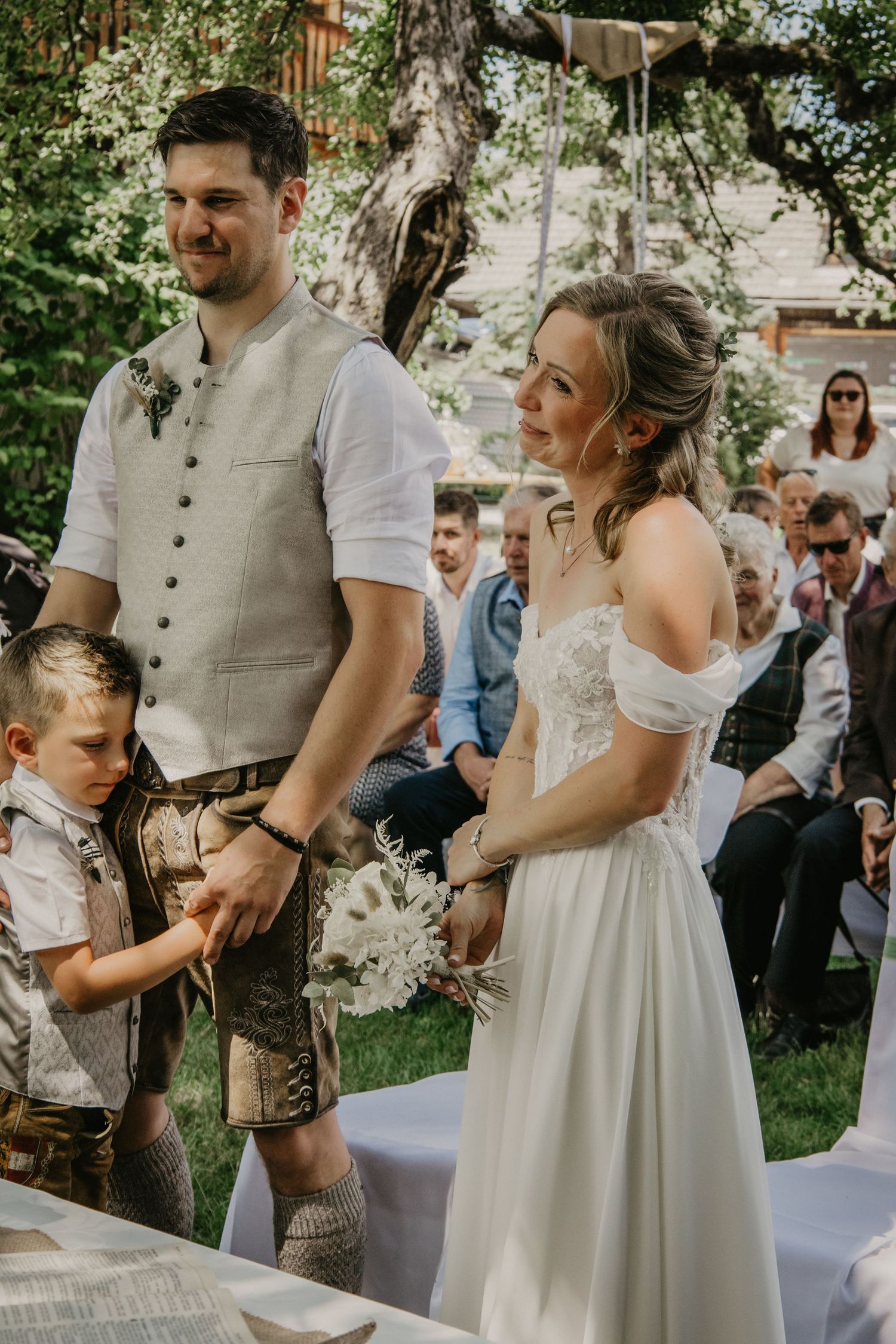 Bride and groom standing at outdoor wedding ceremony with young boy beside them holding hands.