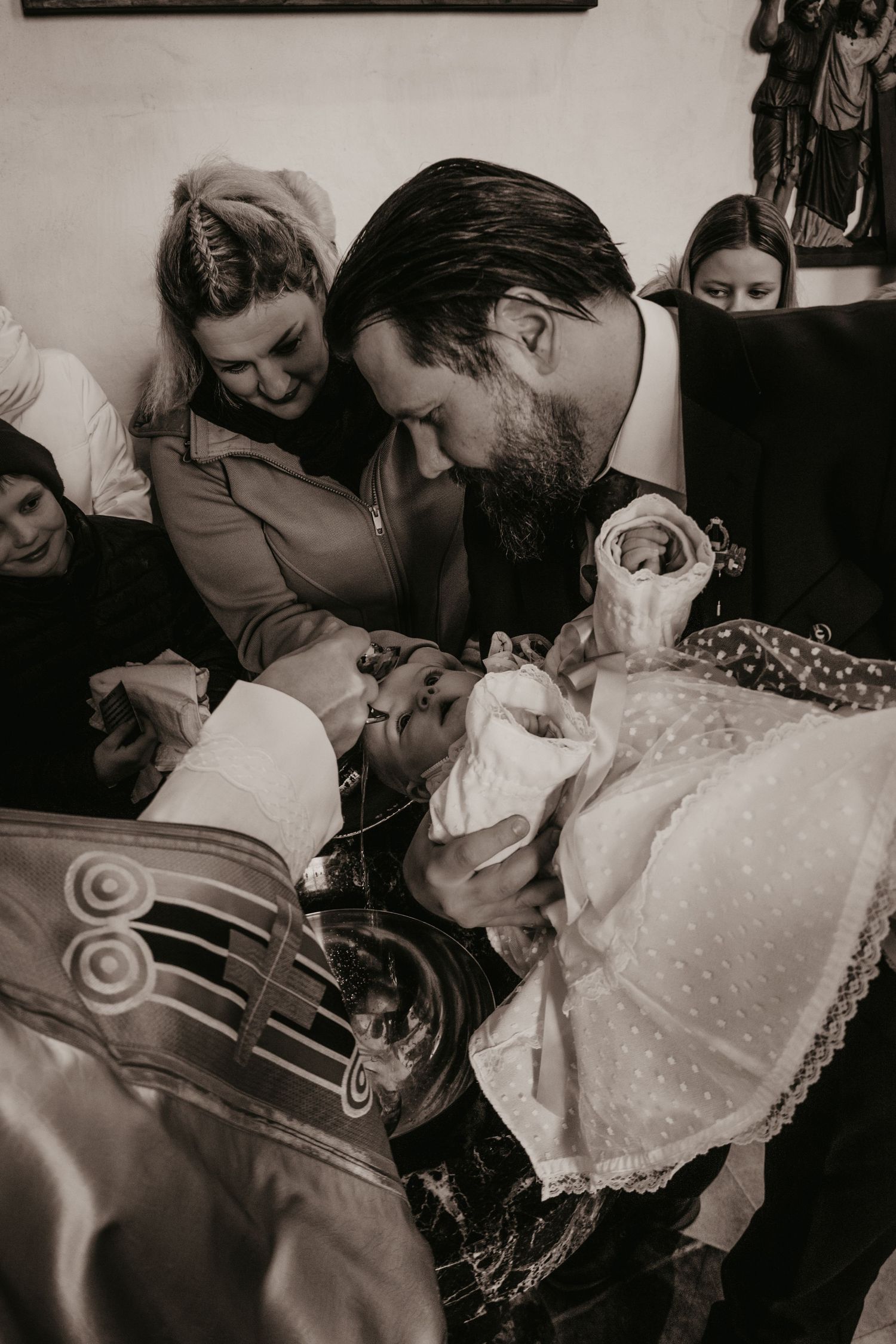 Parents and family lean over a baby in a lace christening gown during a black and white baptism ceremony at the font.