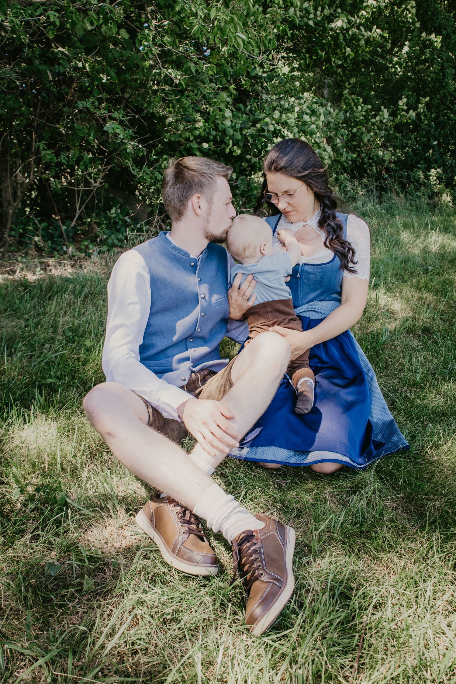 Young family sitting on grass, parents in blue outfits lovingly attending to their baby boy outdoors.