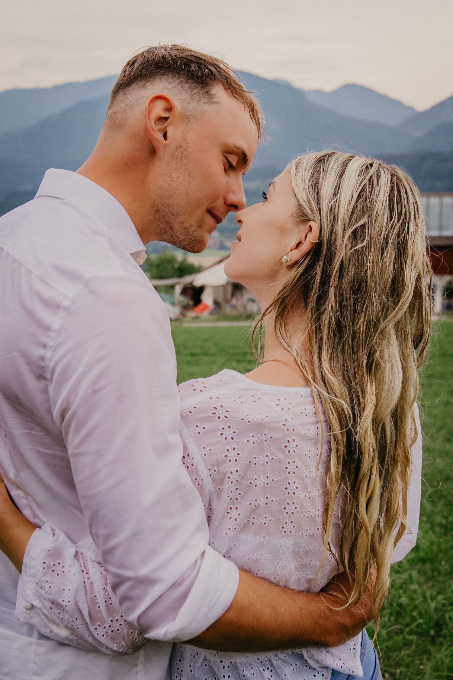 Romantic couple about to kiss outdoors with mountain backdrop, embracing in soft natural light.