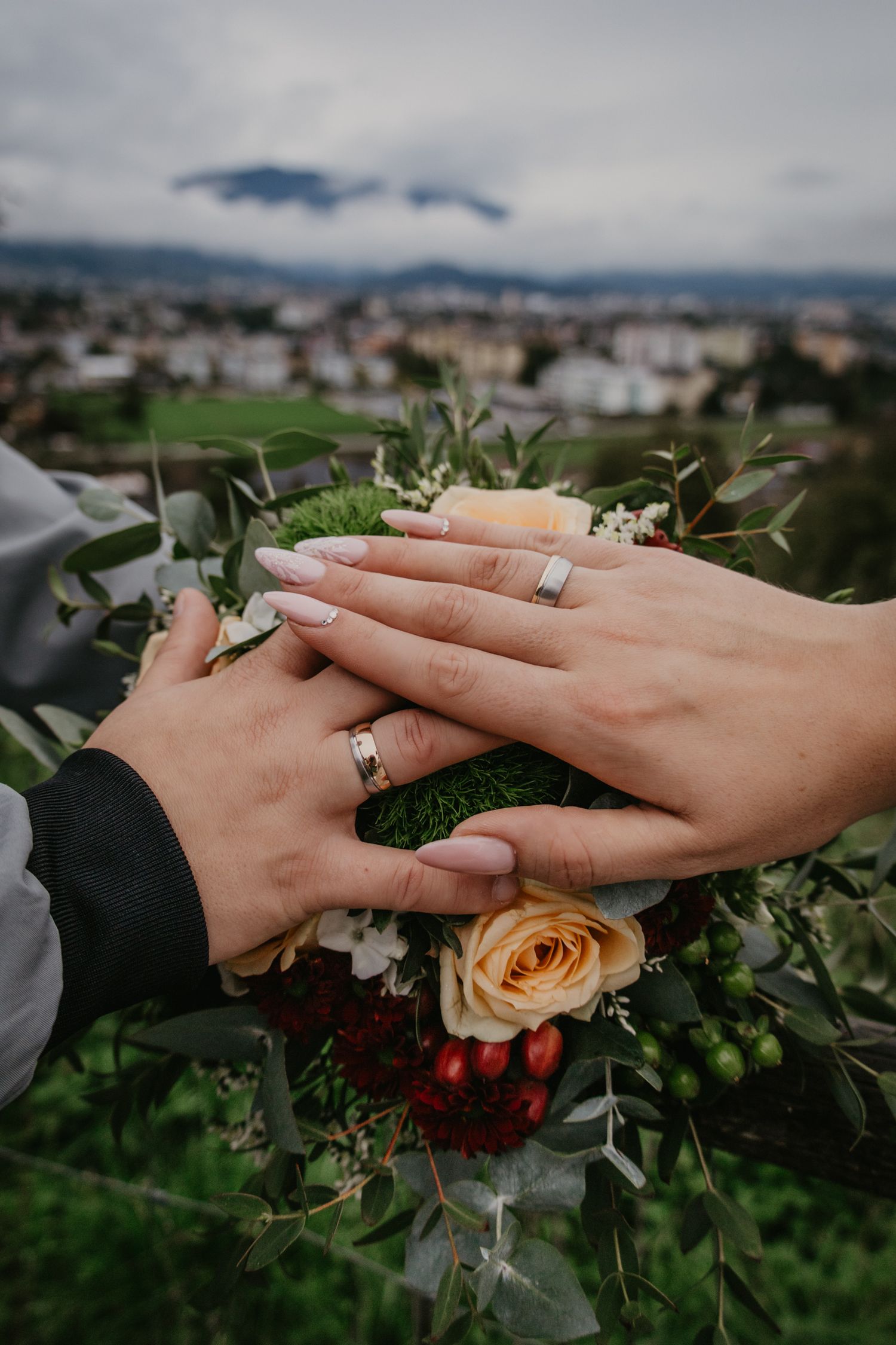Wedding rings and hands displayed with peach roses against scenic mountain backdrop.