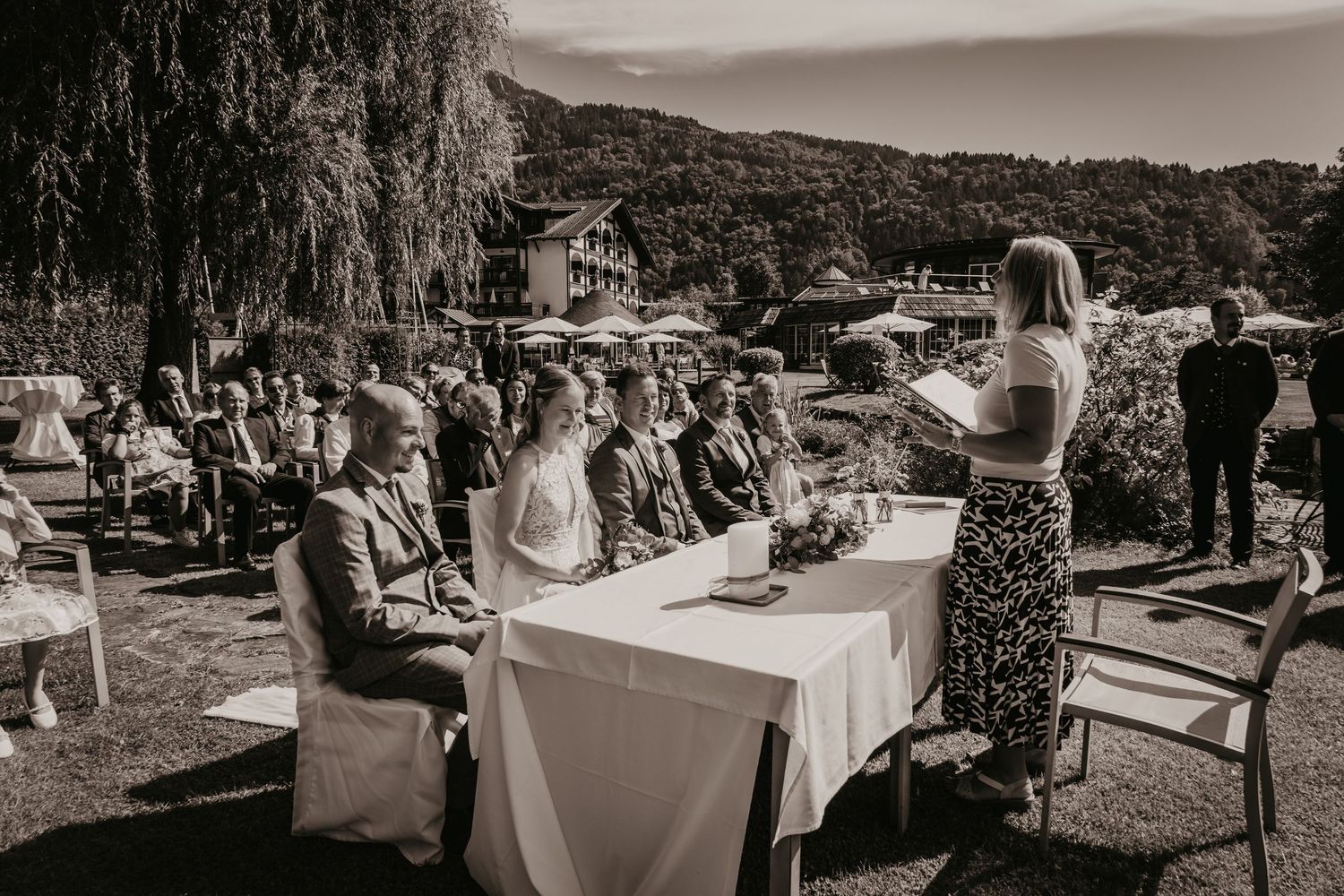 Outdoor wedding reception setup with white tables and chairs under tall trees.