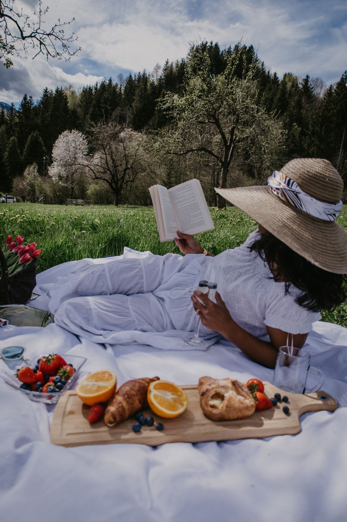 Rustic picnic spread with wooden serving board, fruit, and flowers on white blanket.