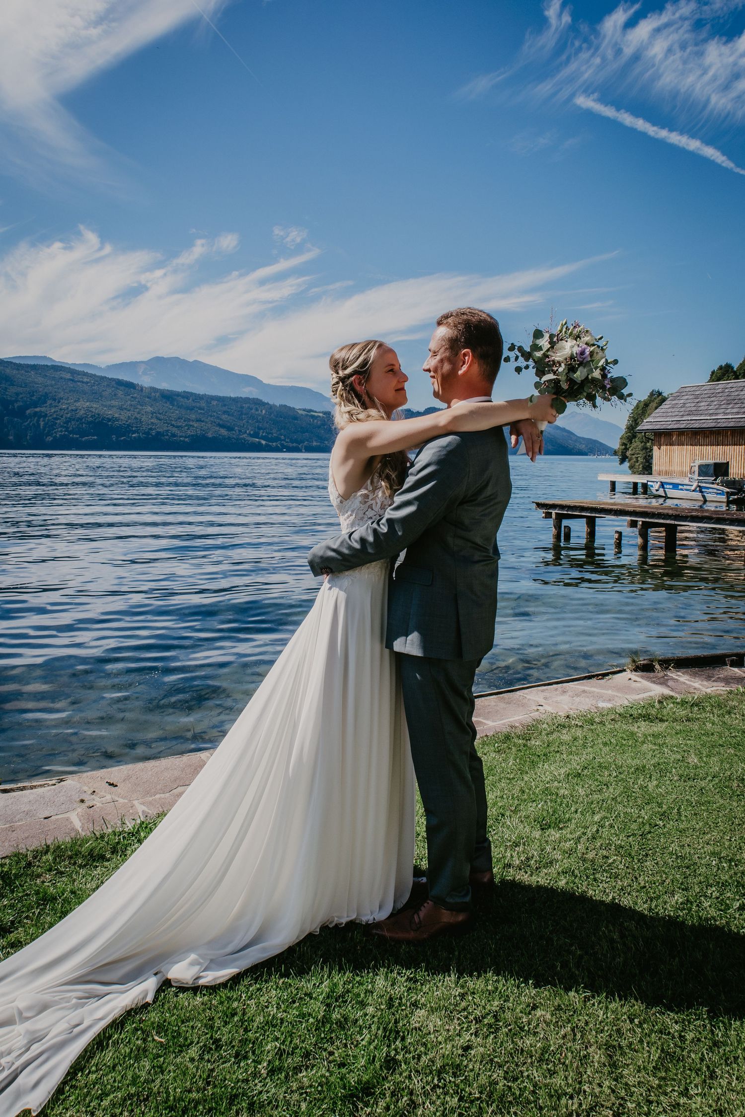 Newlyweds embrace on grassy shoreline with lake and mountain vista behind them.