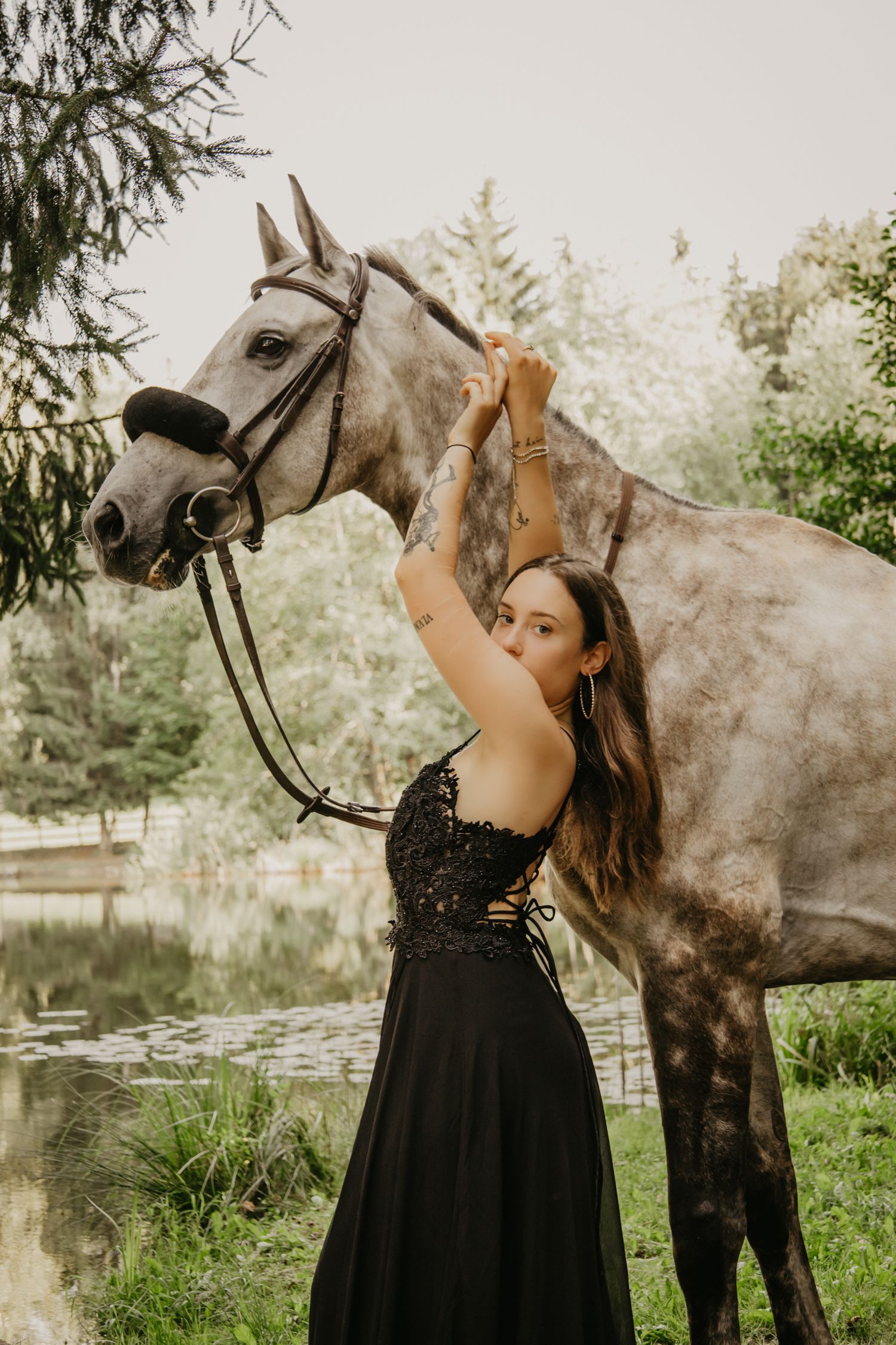A graceful pose with arms raised next to a grey horse by a pond.