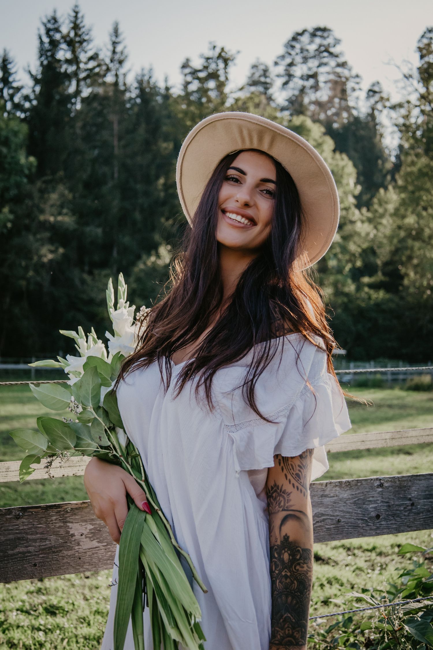 Sun-drenched portrait with white flowers, straw hat, and white dress in country setting.