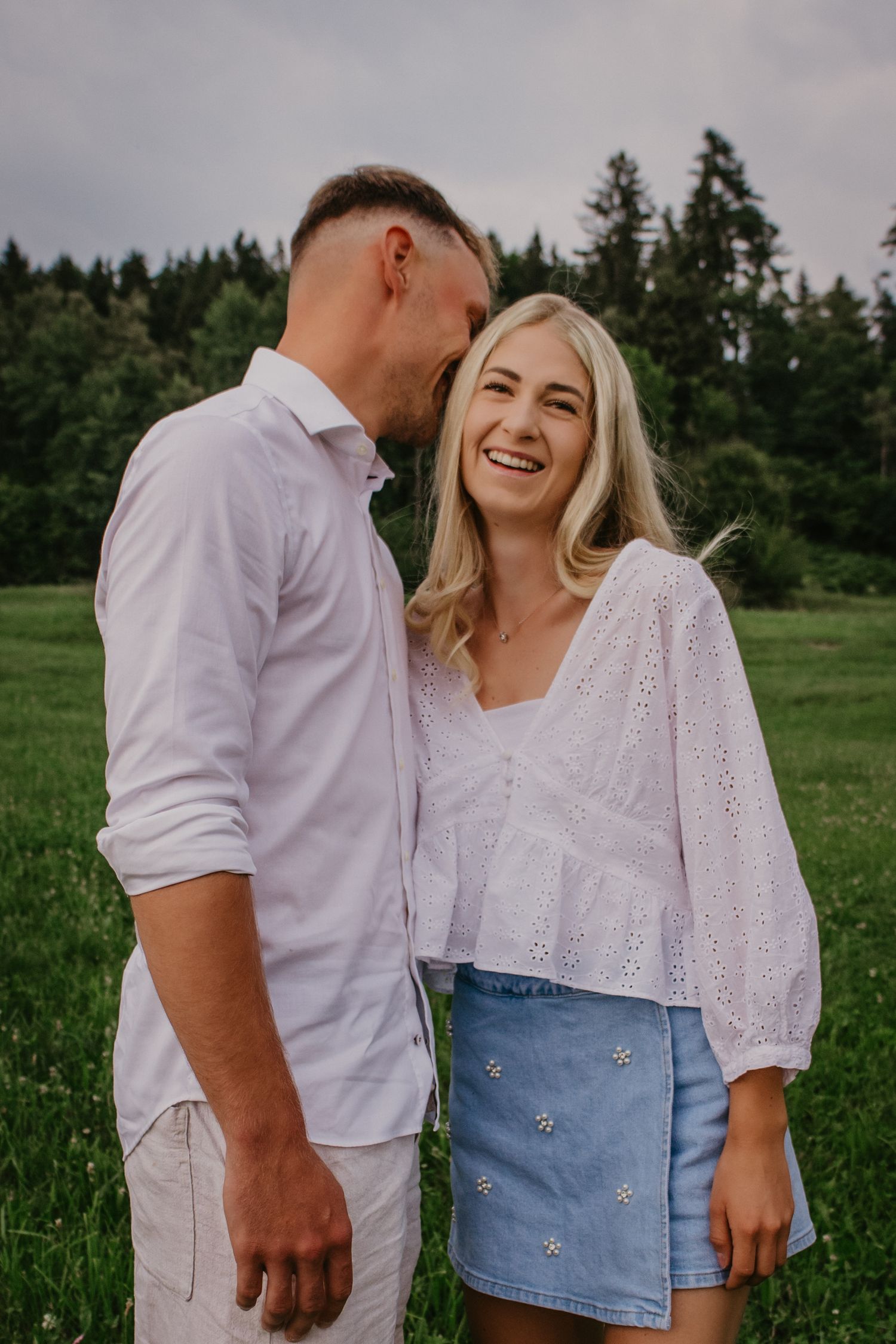 A smiling blonde woman in a white blouse and denim skirt stands with a man in a green field.