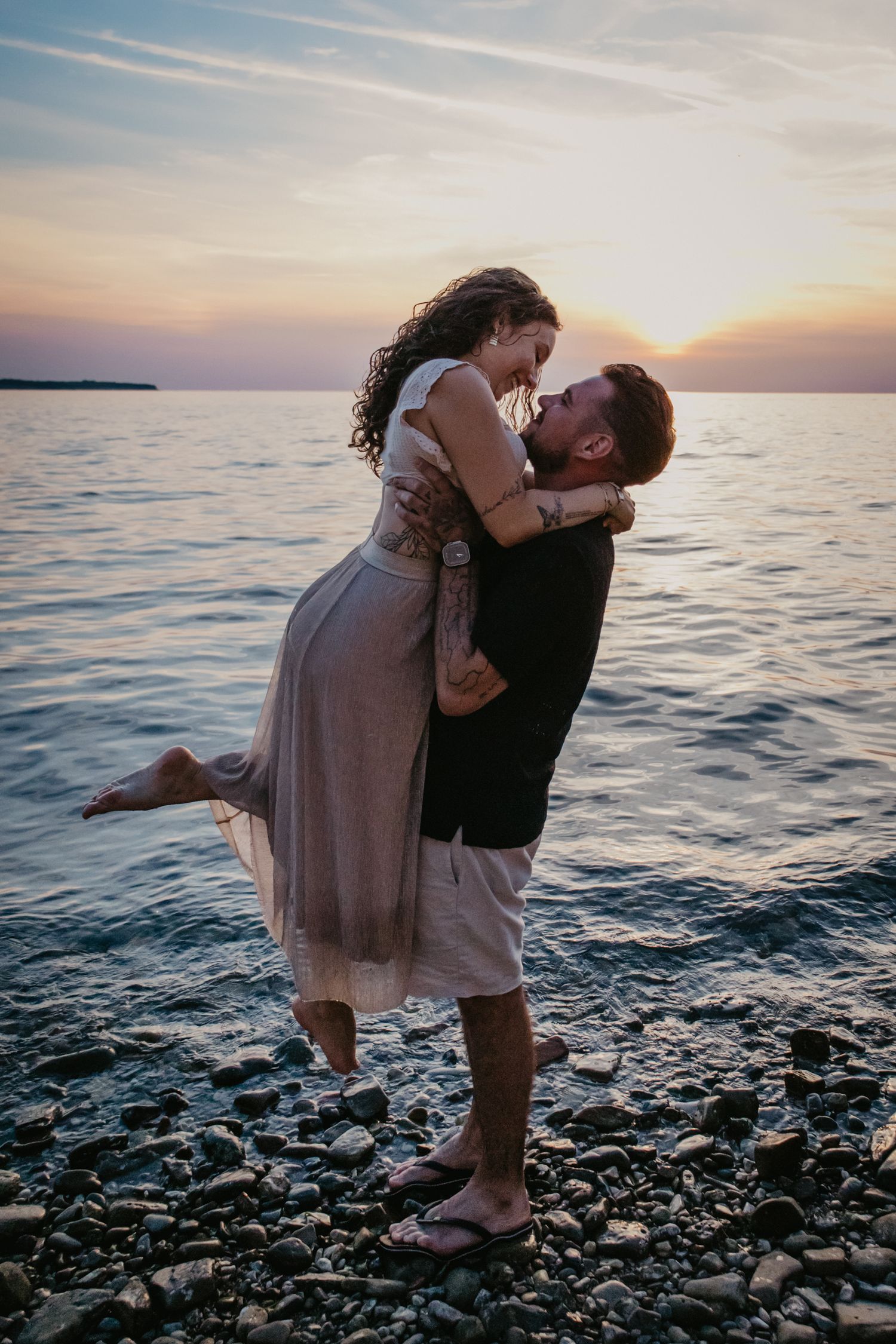 Two people share a romantic embrace on a rocky beach during sunset.