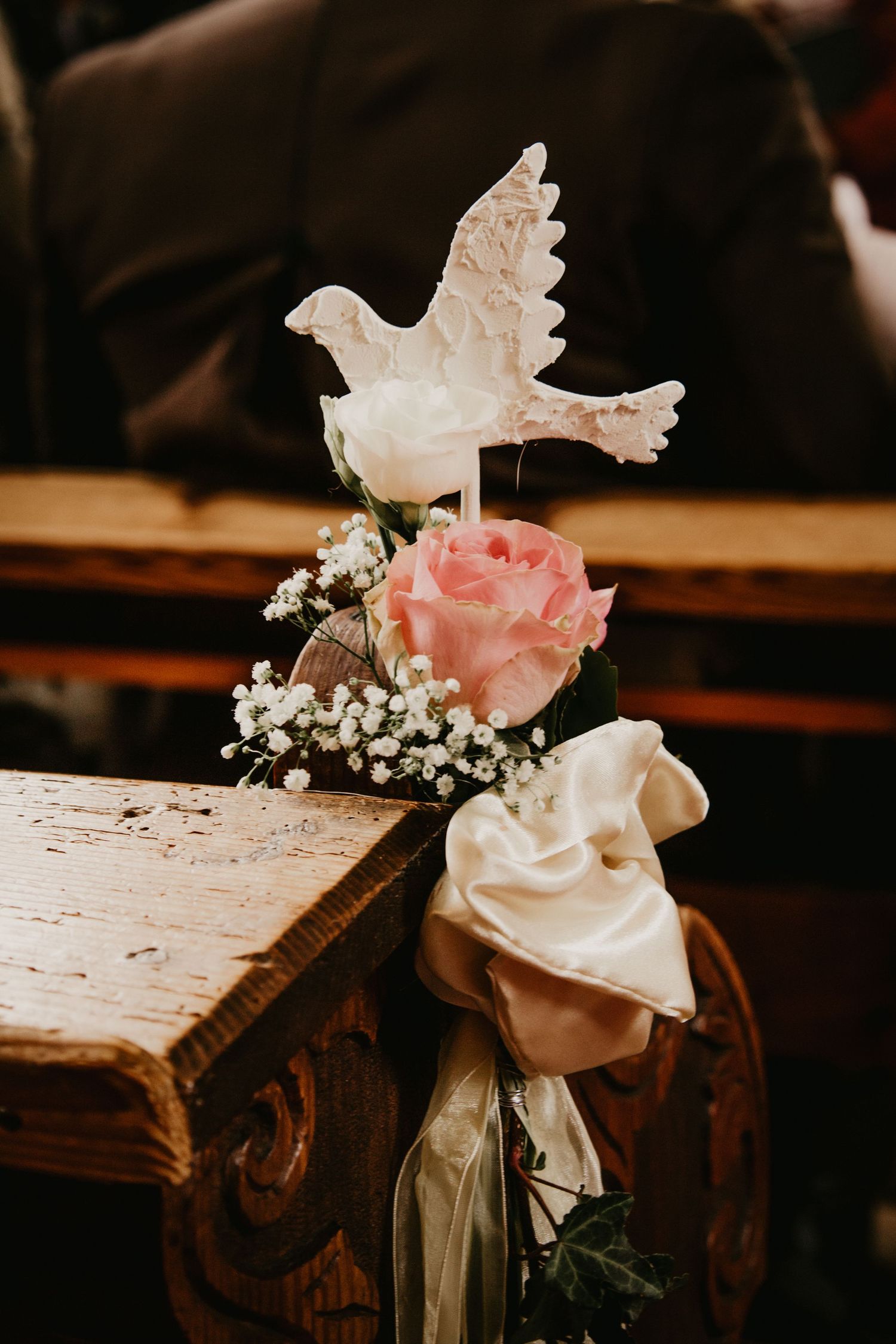 Church pew decorated with pink and white roses, baby's breath, a white dove ornament, and satin ribbon for a wedding ceremony.
