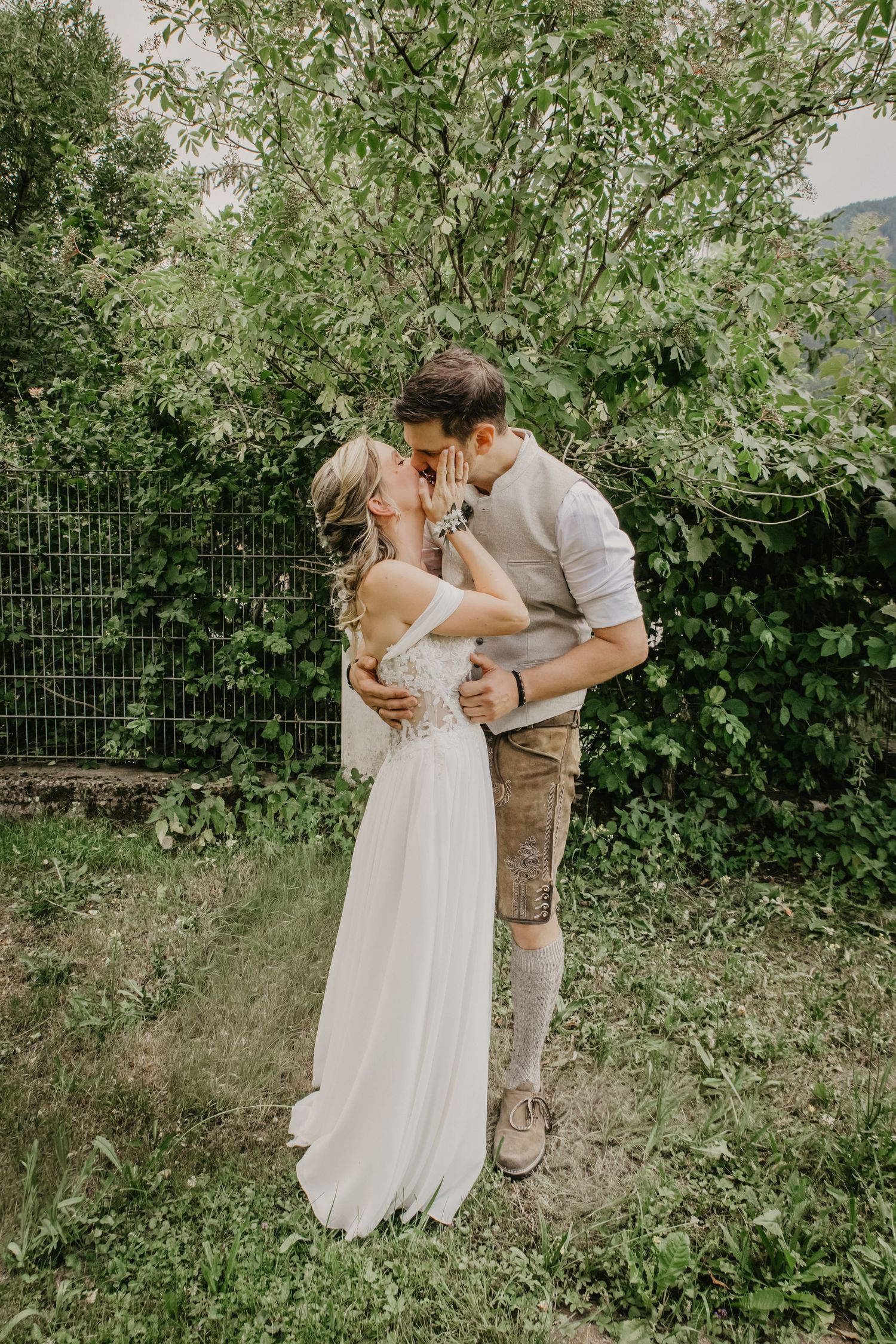 A couple shares a romantic kiss in a lush garden, bride in white gown, groom in traditional Bavarian lederhosen.