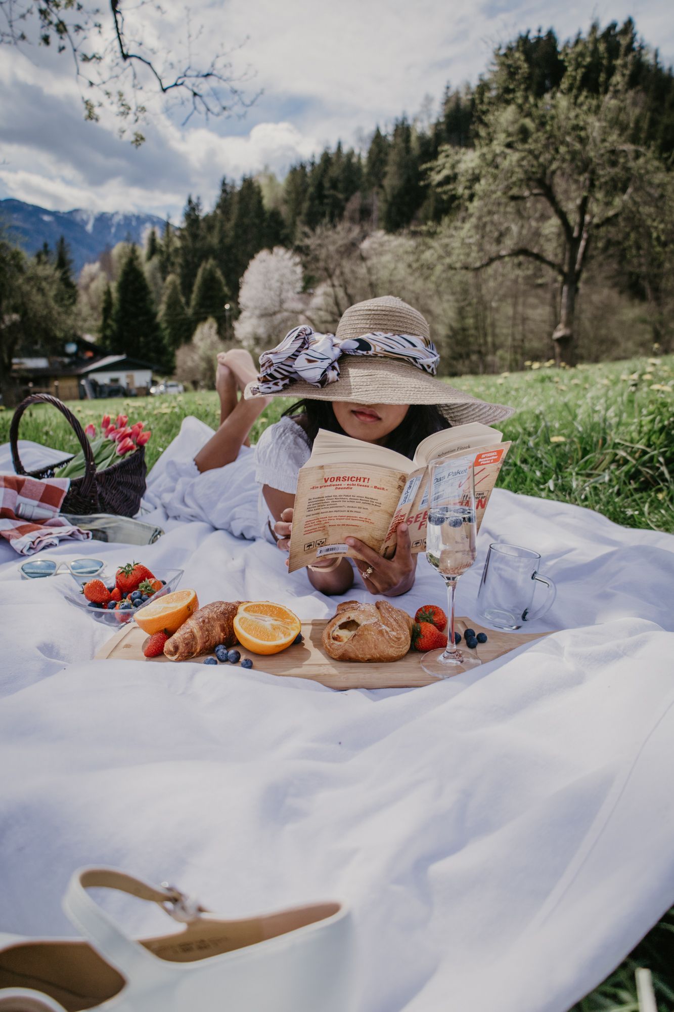 A picturesque outdoor picnic spread on a white blanket with fresh fruit, bread, and snacks against a mountain backdrop.