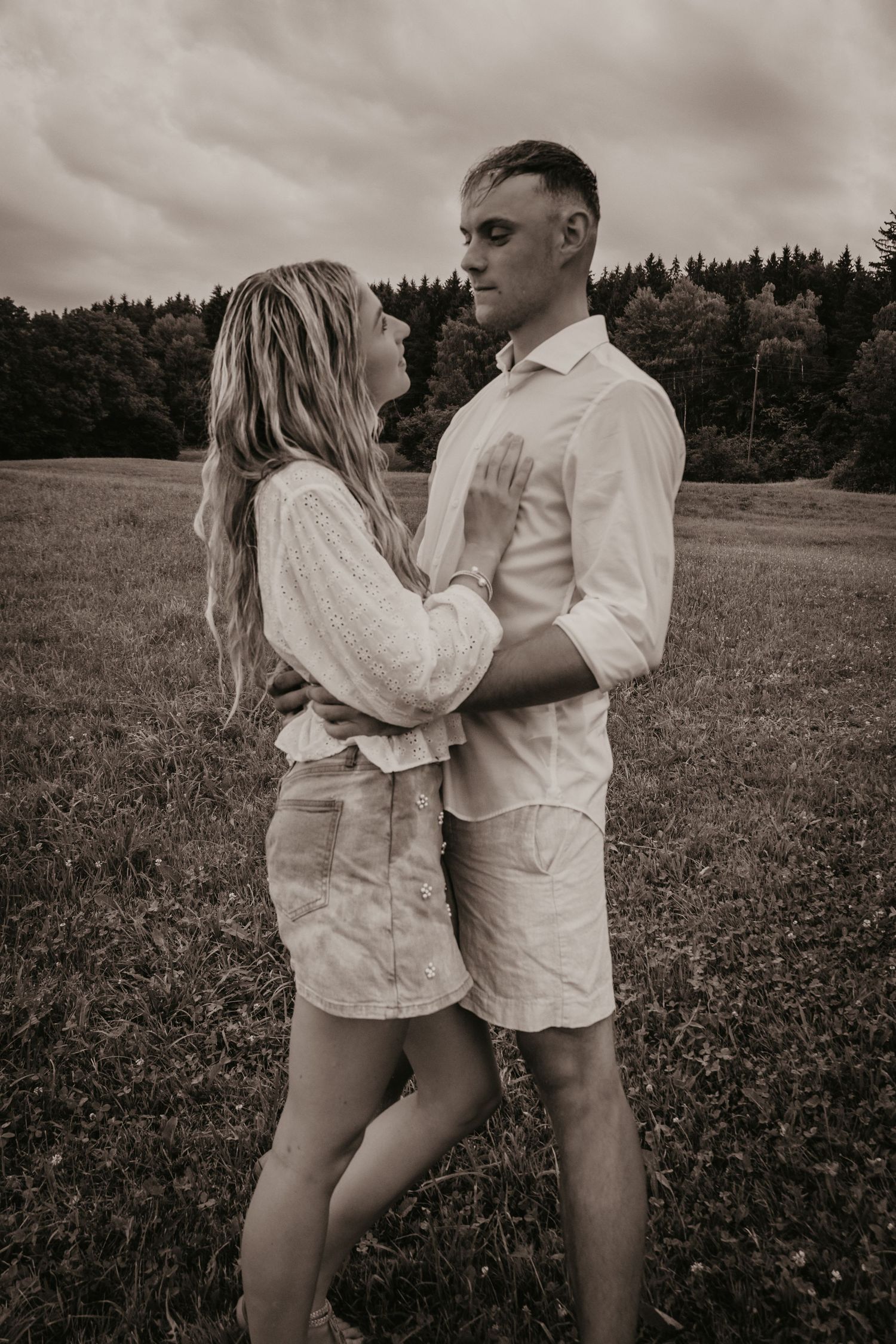 Romantic couple embracing in sepia-toned outdoor portrait with overcast sky and forest backdrop.