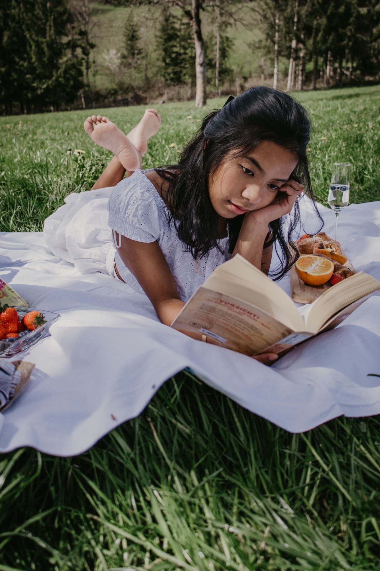 A peaceful outdoor reading session on a white blanket with fresh fruit and snacks in a grassy field.