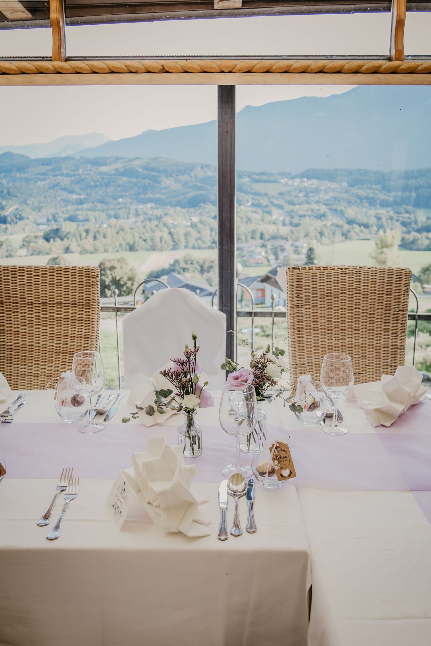 Elegant wedding reception table with wicker chairs and mountain views through large windows.