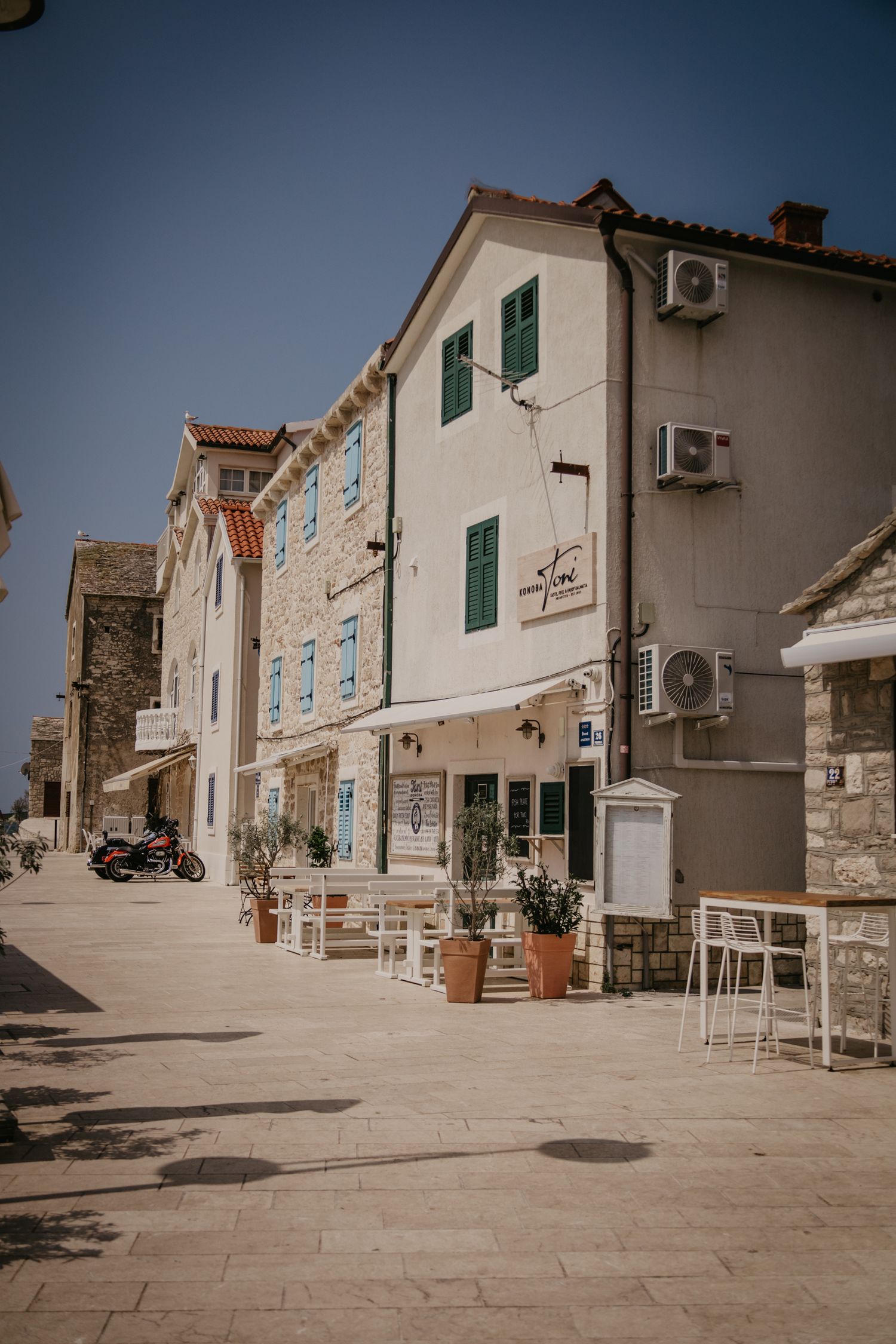 Traditional white stone buildings line a sunny Mediterranean street with outdoor seating.