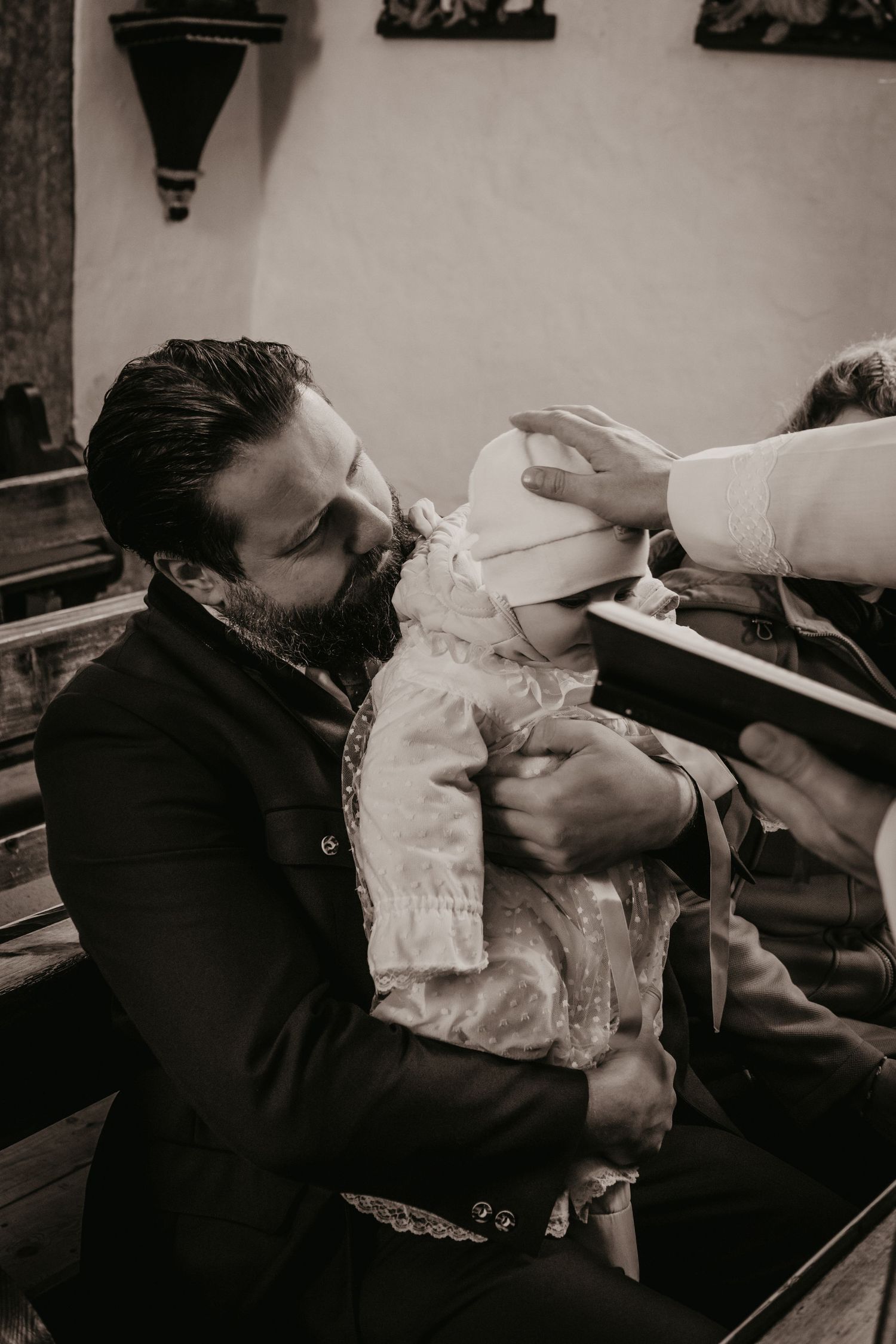 Bearded father in a dark jacket holds his baby in a christening gown while a priest blesses the child's head during a baptism.