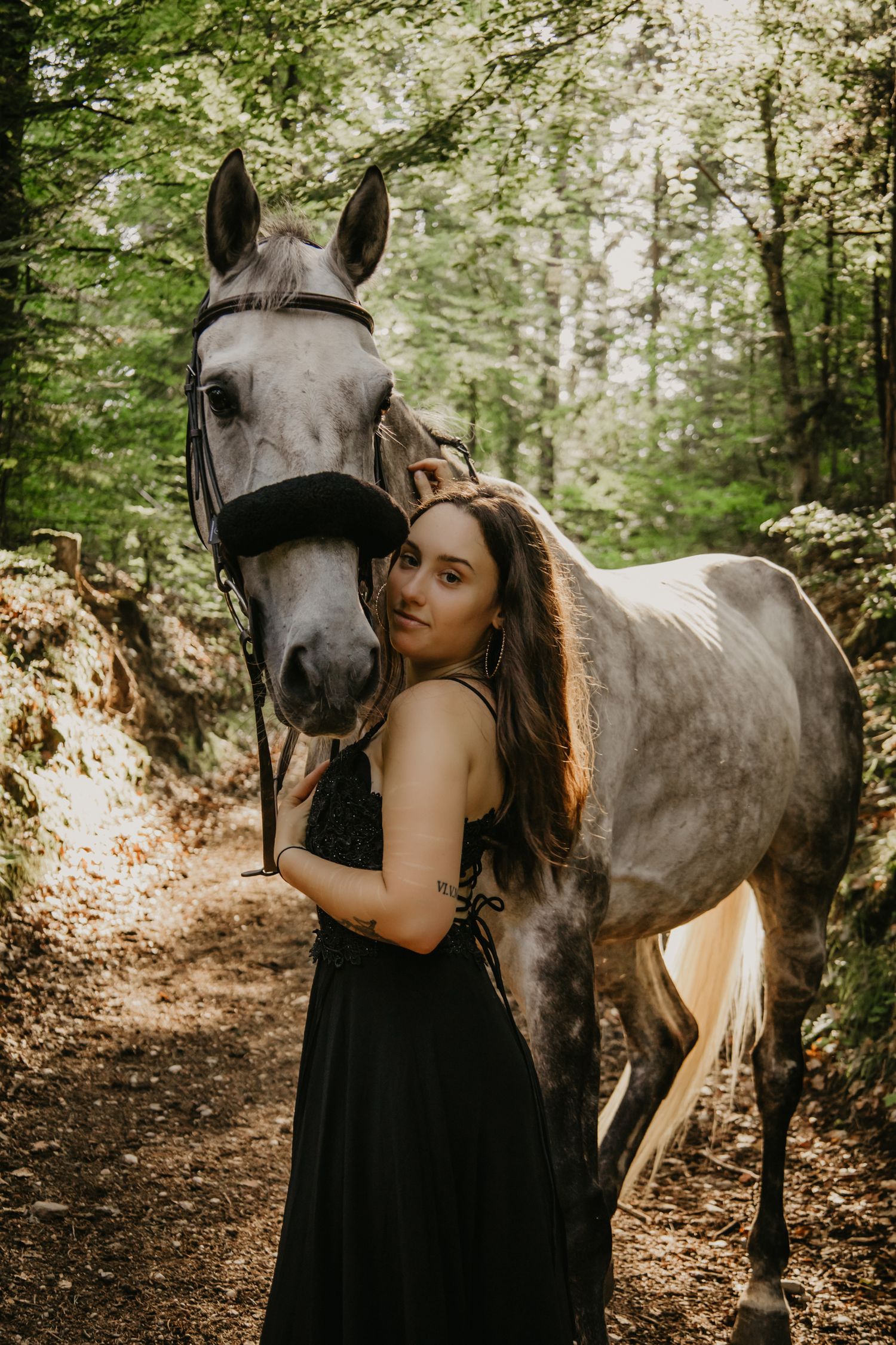 A woman in black dress sharing tender moments with a grey horse in a sunlit forest.