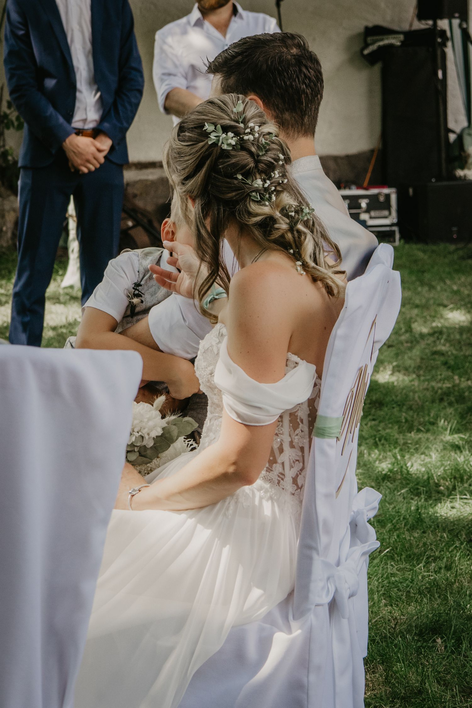 Bride in white off-shoulder gown with floral hair accessory seated outdoors at wedding ceremony.