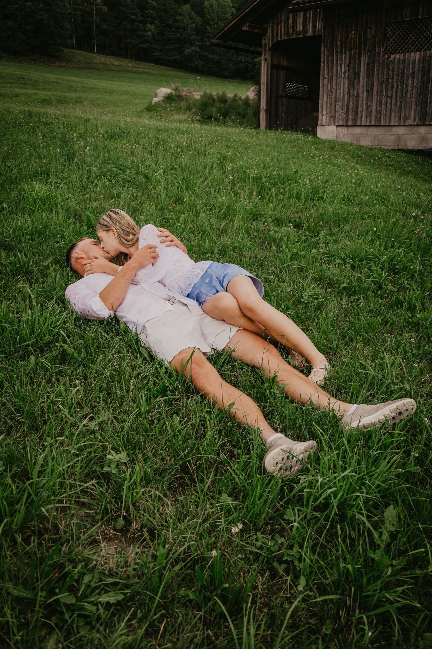 Couple lying together on lush green grass near a wooden barn, sharing a tender romantic moment outdoors.