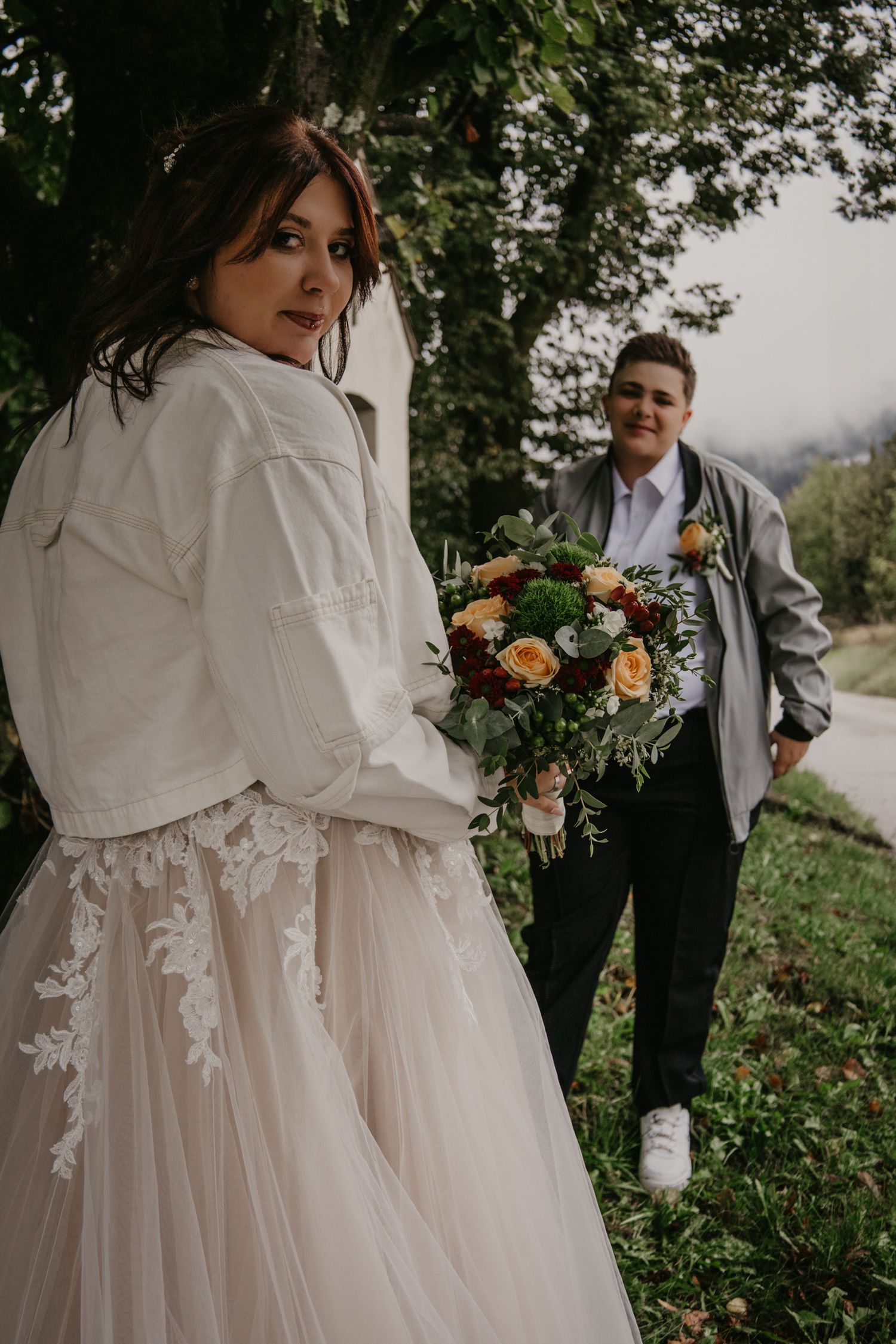 Wedding couple in white dress and grey suit holding floral bouquet outdoors.