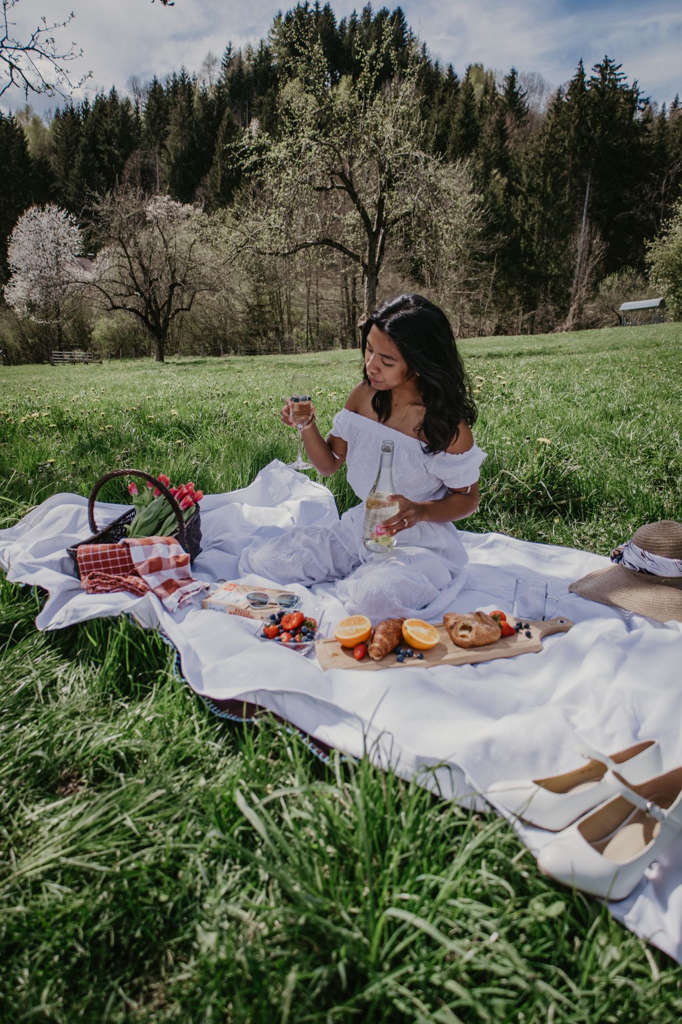 Sunny day picnic spread with food and drinks on white blanket in grassy field.