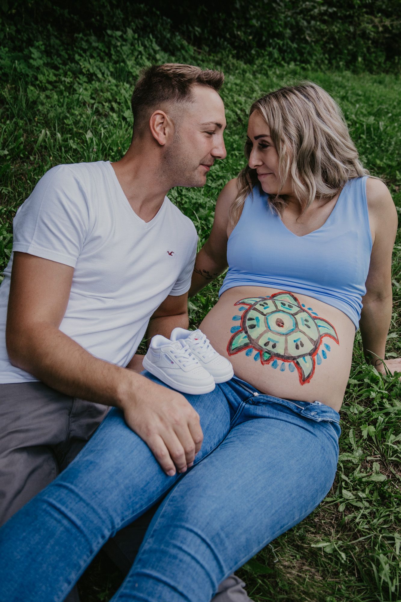 Intimate outdoor maternity photoshoot with a painted belly design against a green natural backdrop.