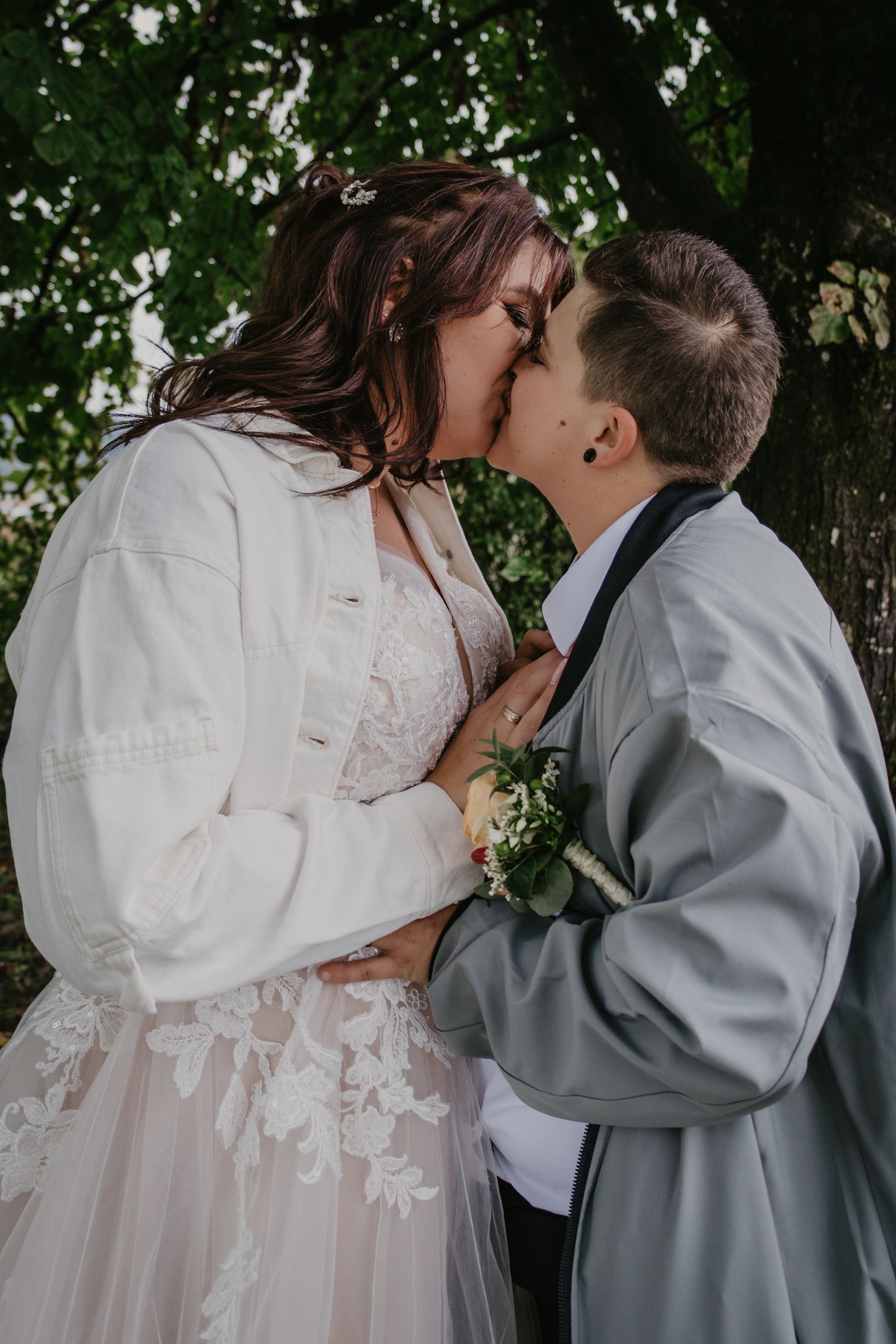Intimate kiss between newlyweds under green tree branches.